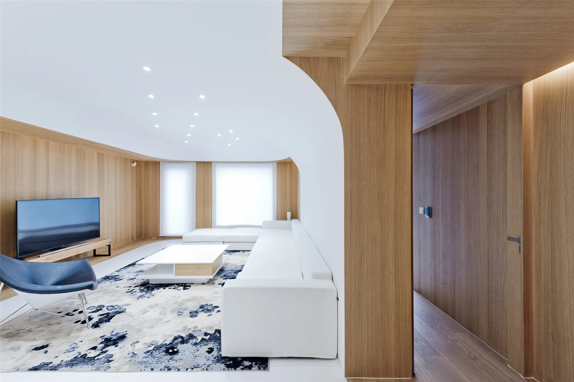 A minimalist living room features a white sectional sofa, a blue accent chair, and custom wood wall paneling with a curved white archway.