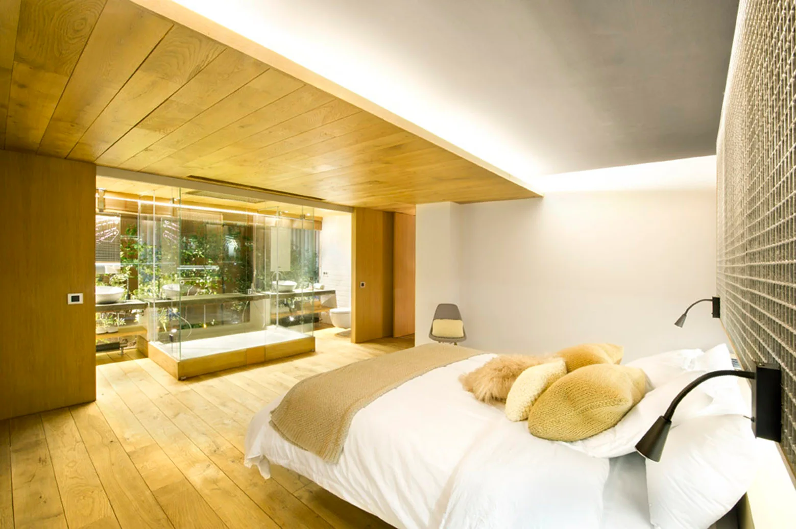 A modern bedroom features a white bed, natural wood plank flooring, a matching wood plank ceiling drop, and a glass-enclosed bathroom beyond.