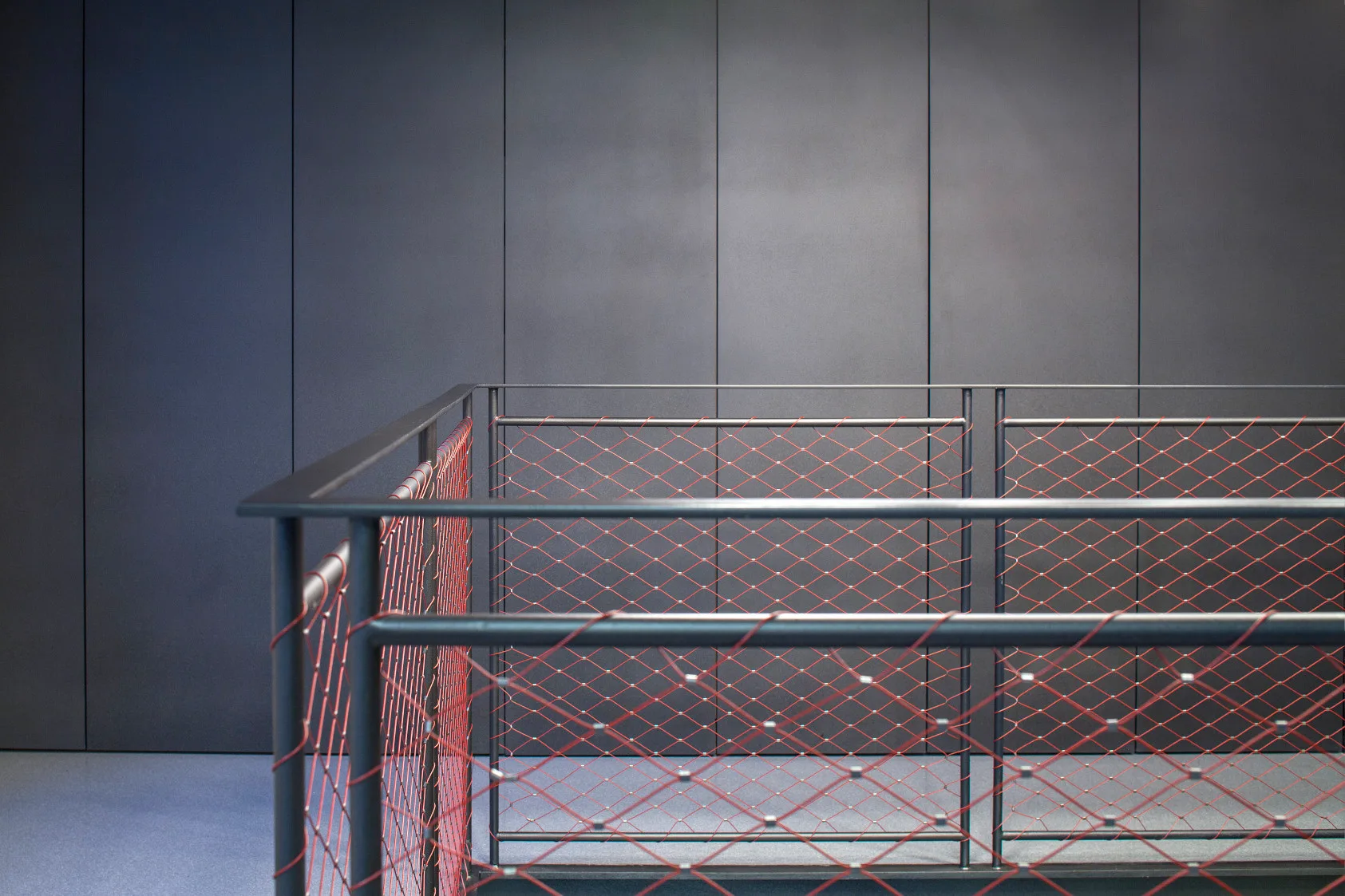 A black metal safety railing featuring a bright red diamond mesh grid stands in front of a dark gray paneled wall.