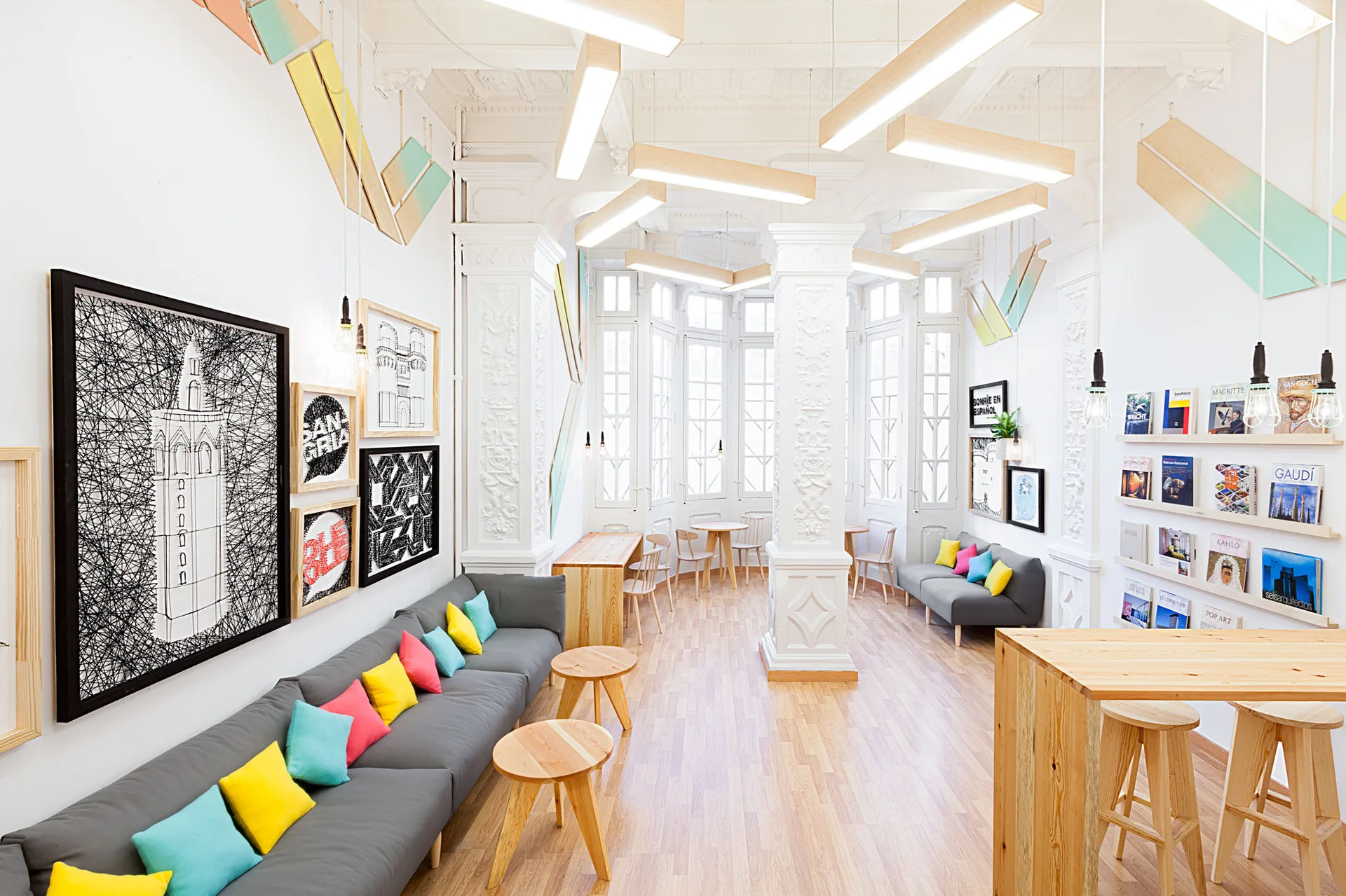A bright lounge features grey sofas with colorful throw pillows, light wood tables, ornate white columns, and linear modern ceiling lights.