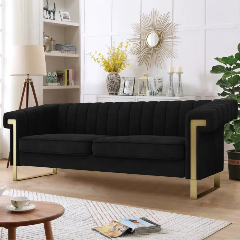 A black channel-tufted sofa with gold metal legs sits in a light-filled living room featuring white wall paneling and a gold sunburst mirror.