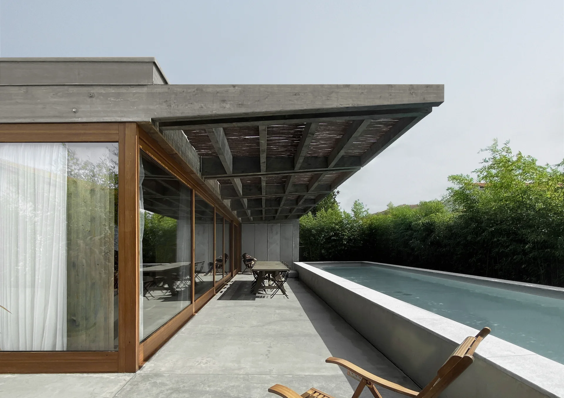 A concrete patio featuring a long rectangular swimming pool, a wood-framed glass sliding door, and tall green bamboo plants along the edge.