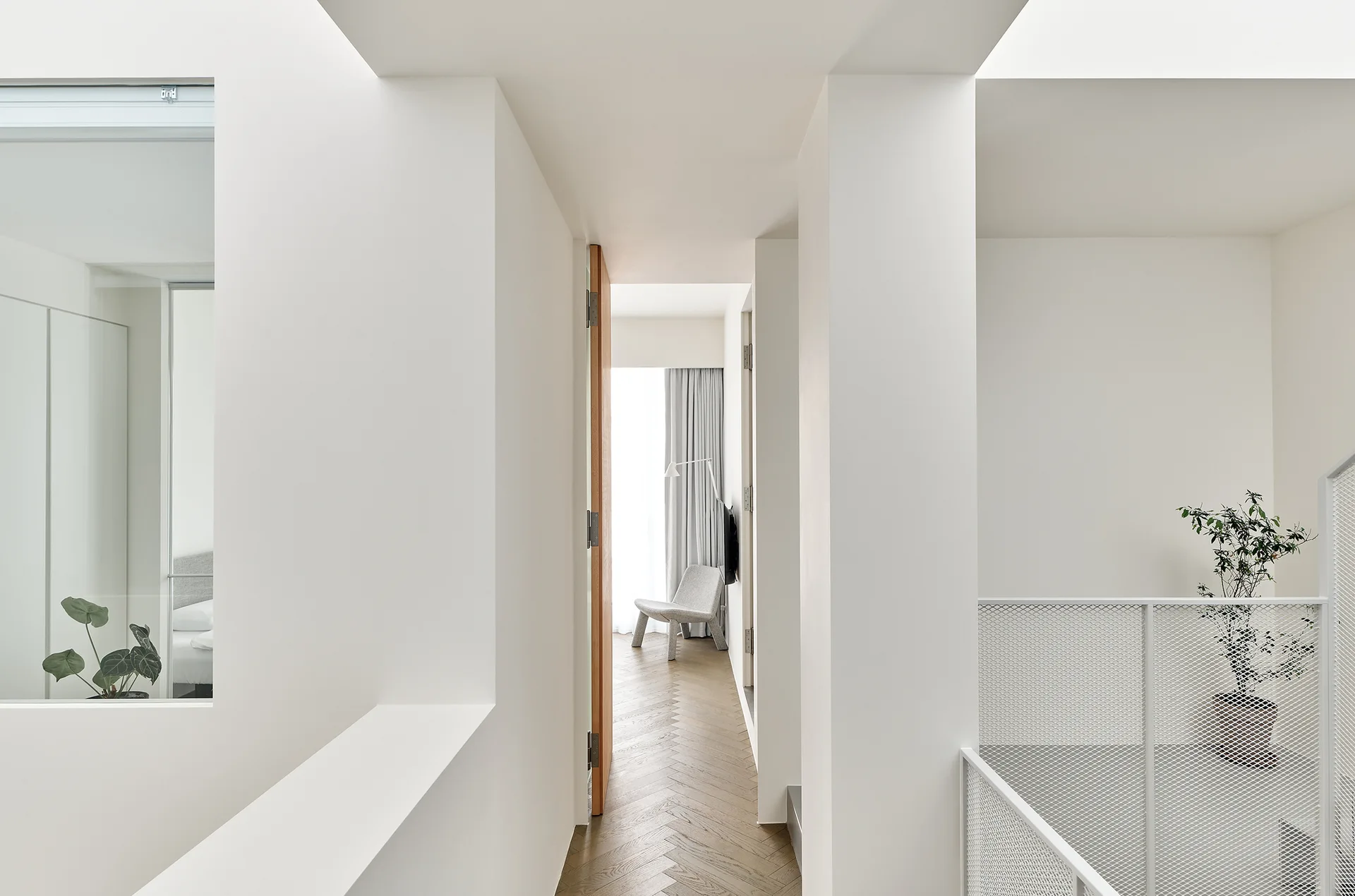A minimalist white hallway featuring herringbone wood flooring, a white metal mesh railing, and a doorway leading to a brightly lit bedroom.