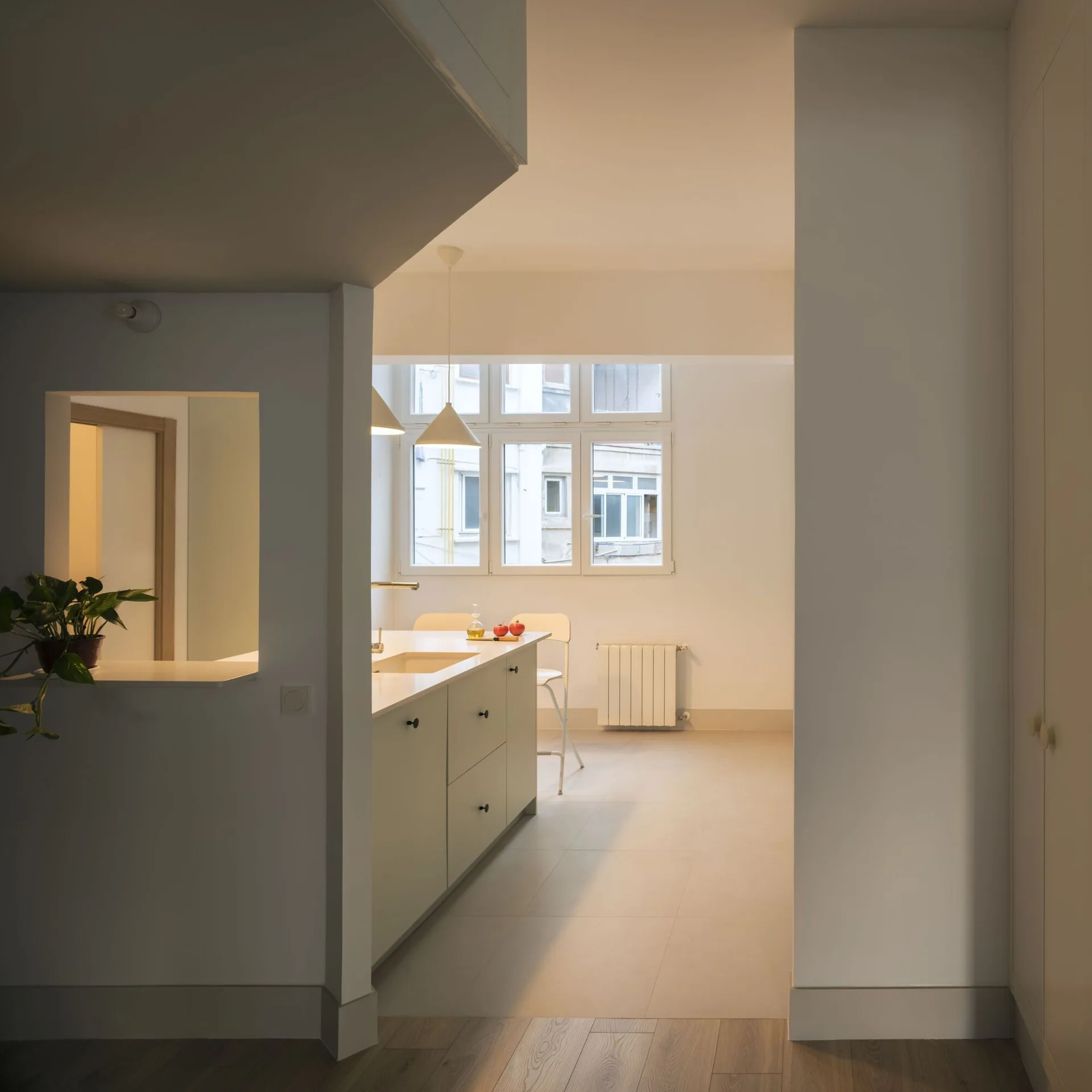 View from a hallway looking into a minimalist off-white kitchen featuring a central island with a sink, pendant lights, and a large back window.