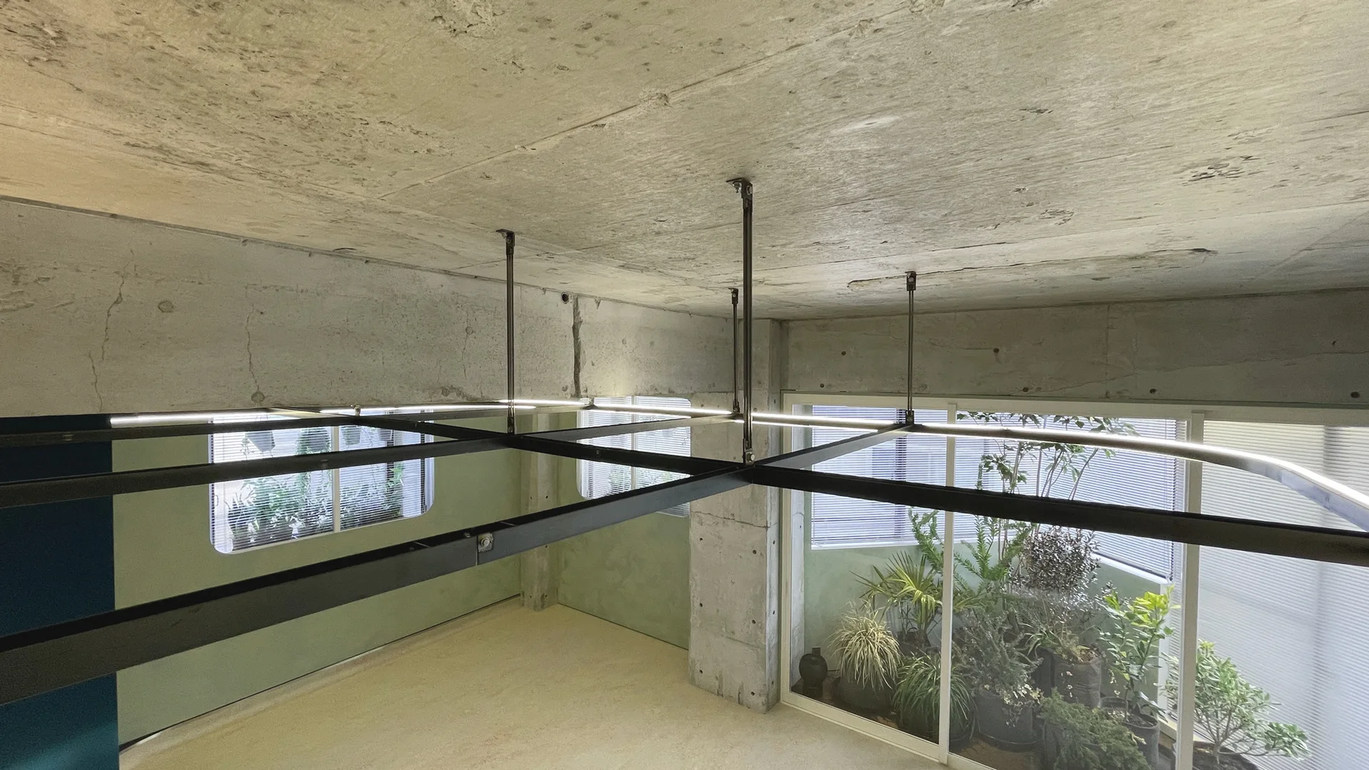 A raw concrete ceiling and pillar frame a suspended black metal lighting grid in an industrial space with sage green walls and courtyard views.
