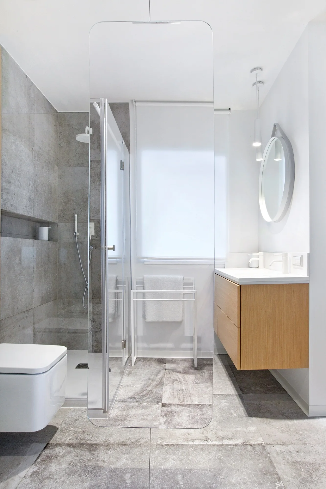 A modern bathroom featuring a floating wood vanity, round mirror, white wall-mounted toilet, and a glass walk-in shower with grey tiles.