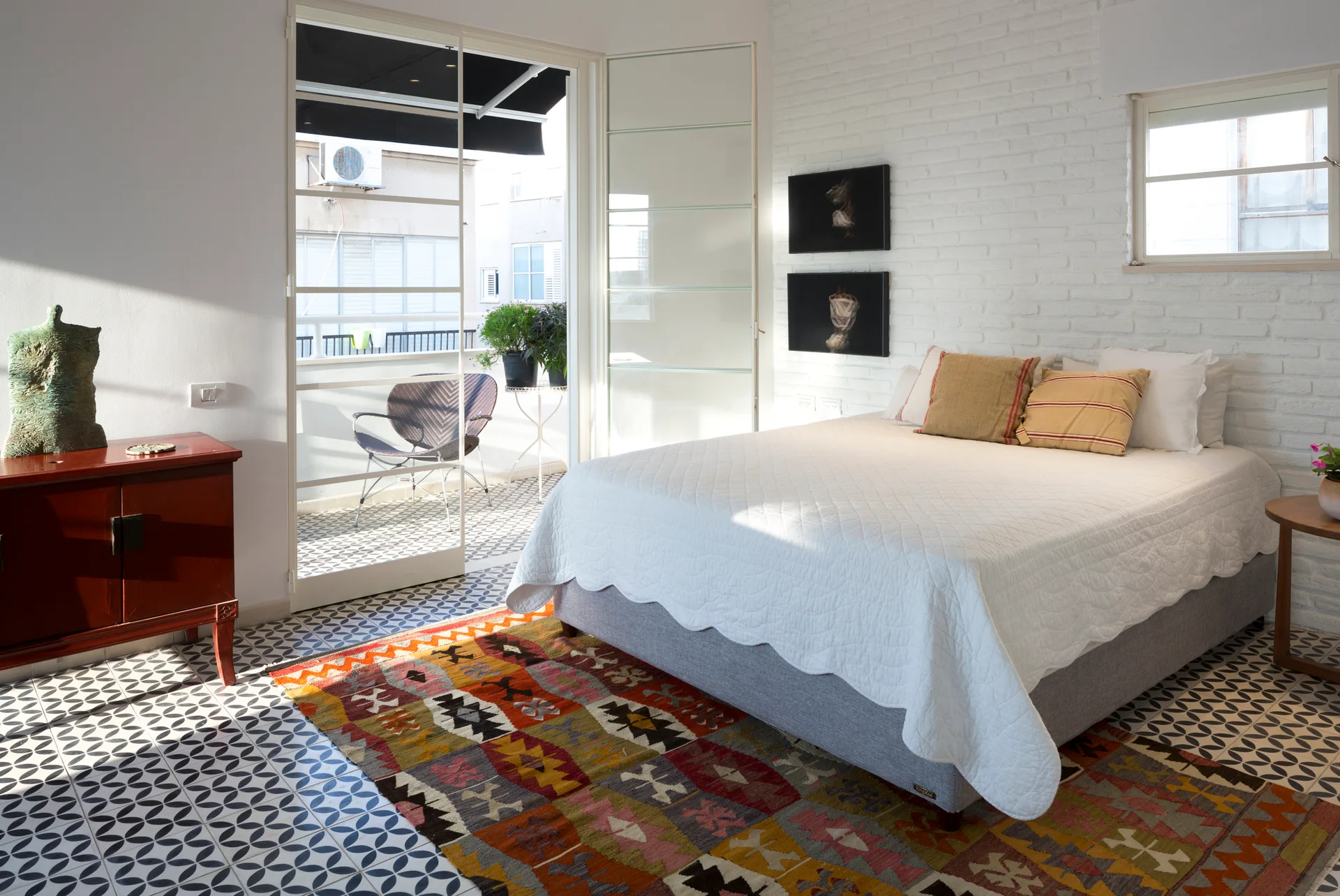 A bright bedroom featuring a white painted brick wall, patterned tile flooring, and a bed with a white quilt next to an open balcony door.