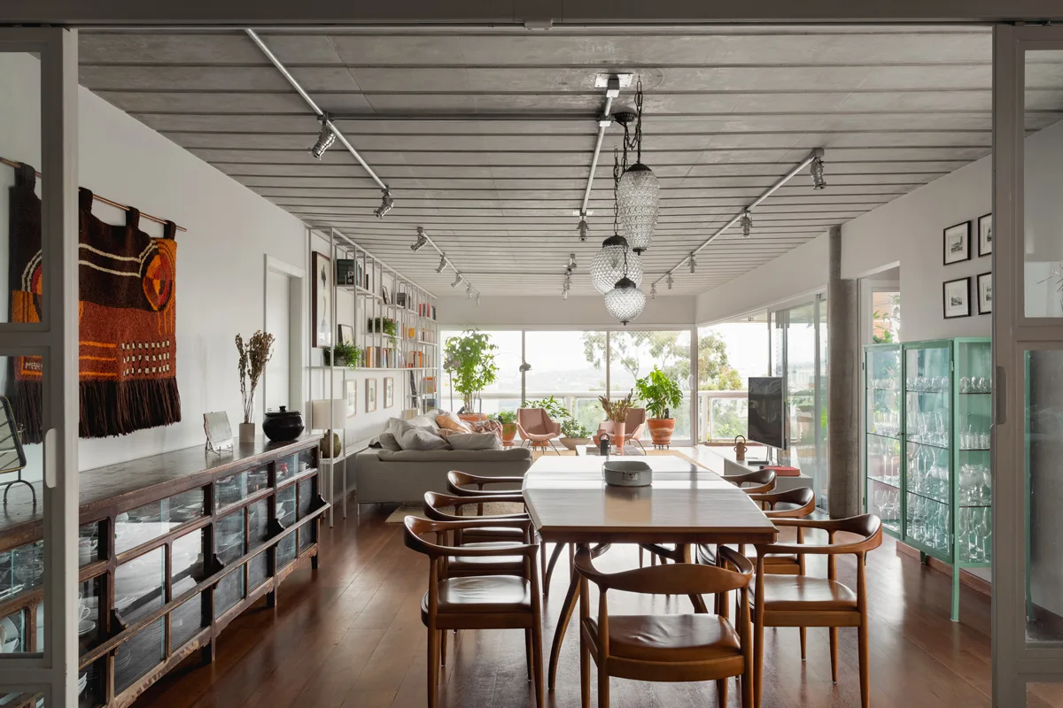 An open-plan dining room features a wooden table and chairs under a concrete ceiling with textured glass pendant lights and a grey sofa in the background.