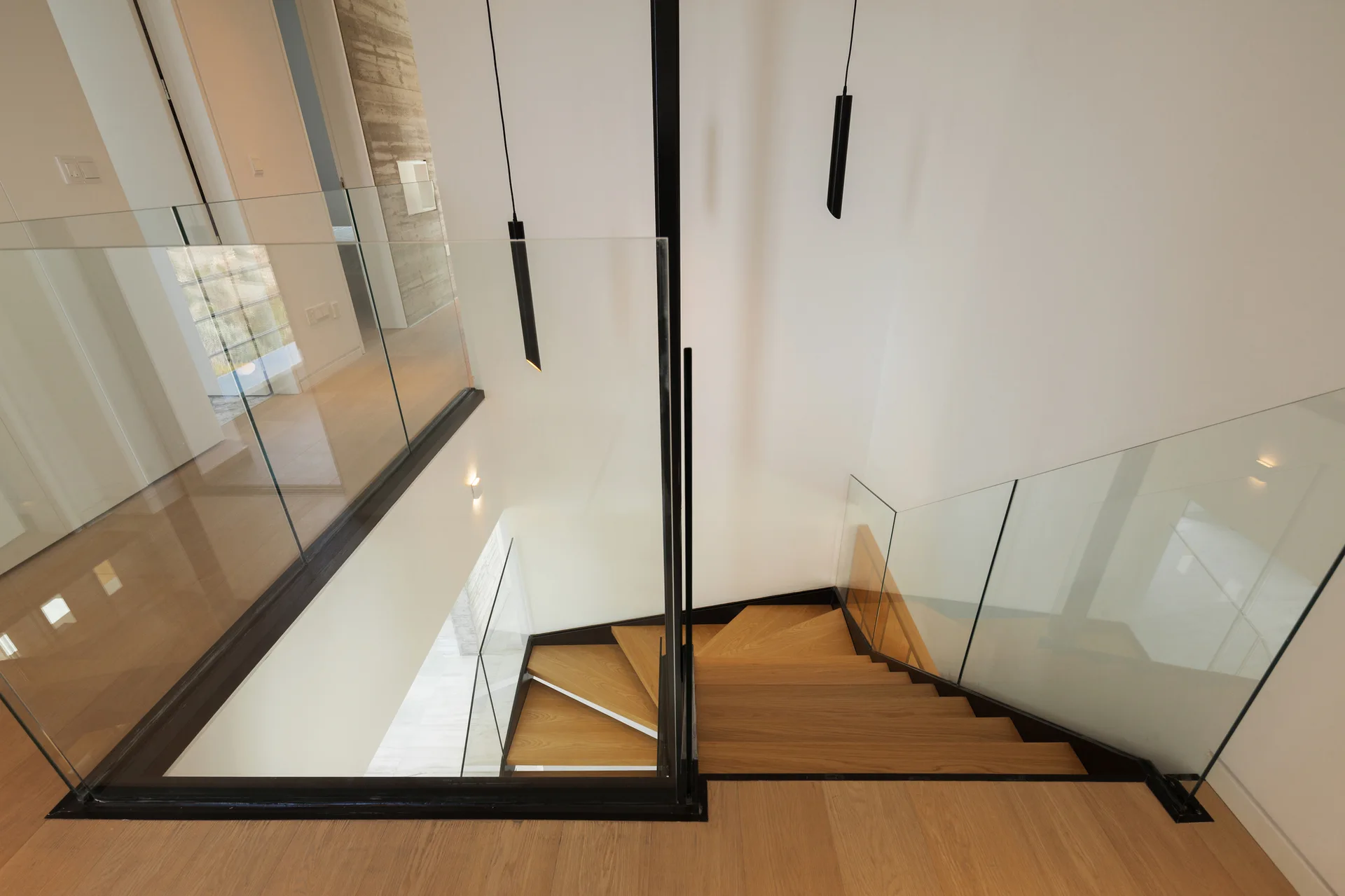 Looking down a modern wooden staircase featuring frameless glass railings and slim black pendant lights hanging in the open stairwell.