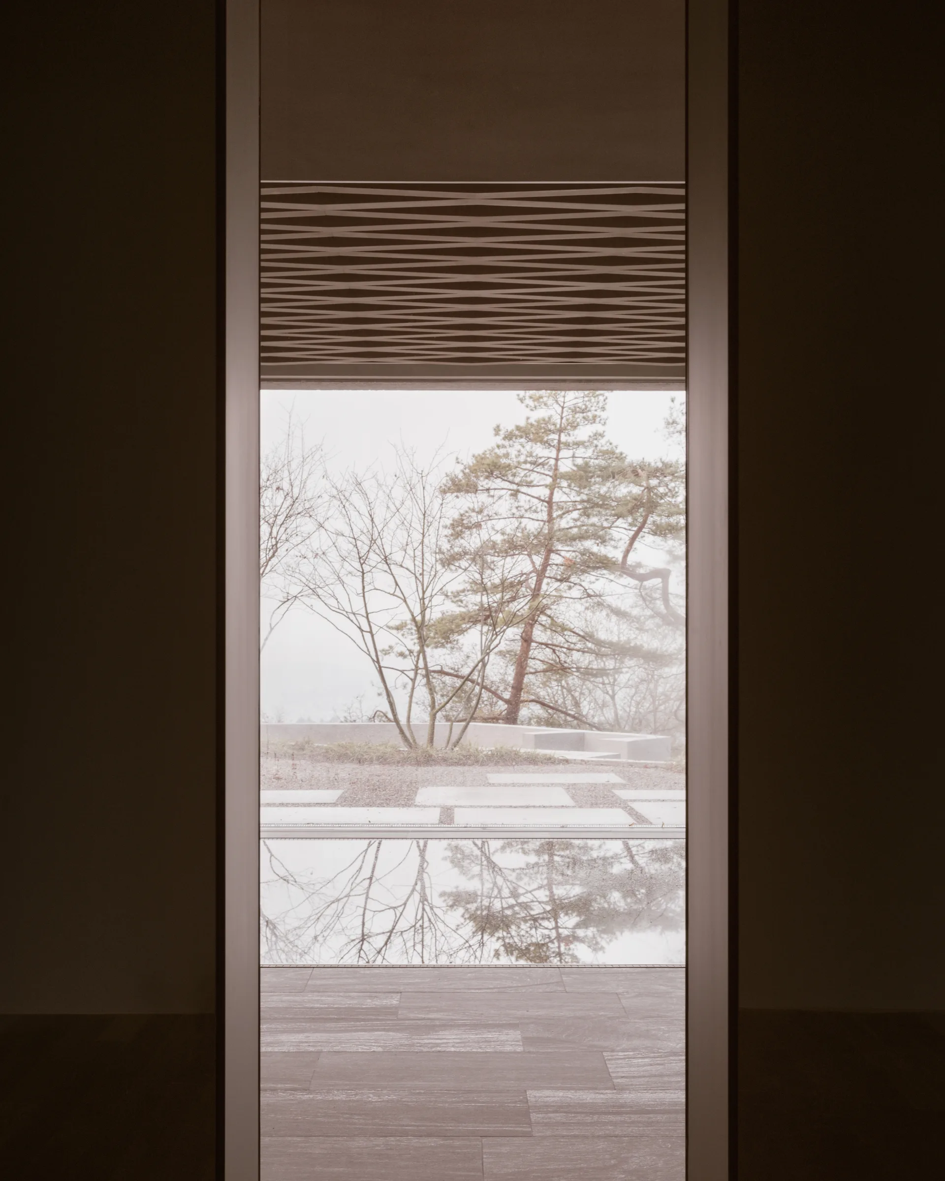 A tall rectangular doorway frames a view of an outdoor gravel courtyard with stone pavers, a reflecting pool, and trees under a foggy sky.