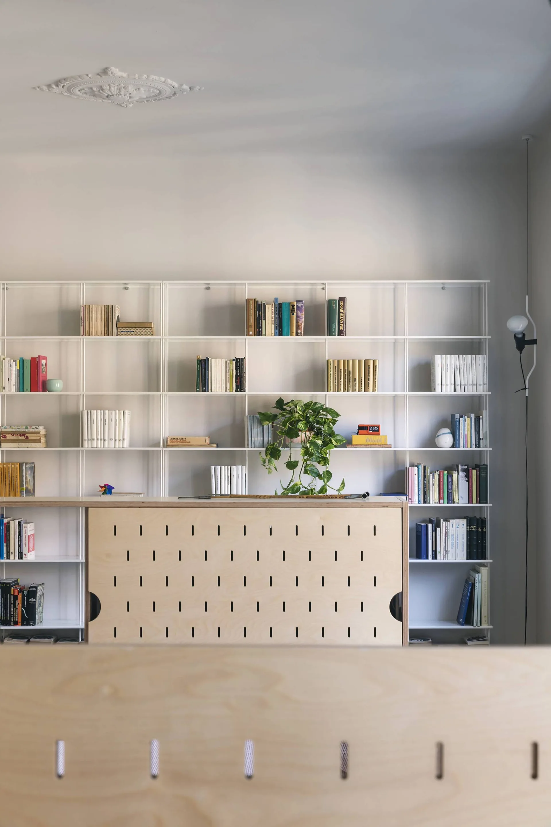 A white metal grid shelving unit filled with books stands behind a light wood plywood desk with vertical slotted details.