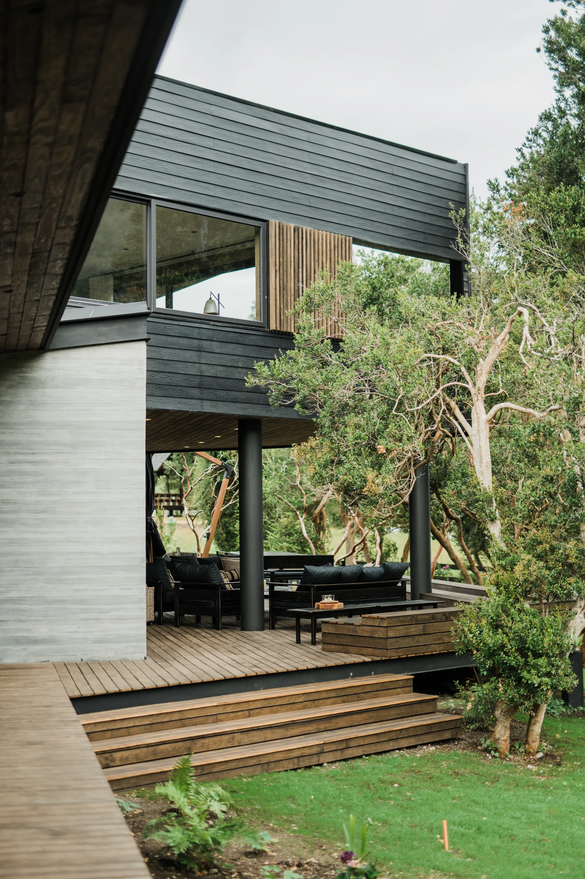 A modern home exterior with charcoal horizontal wood siding, a wooden plank deck with black patio furniture, and steps leading to a green lawn.