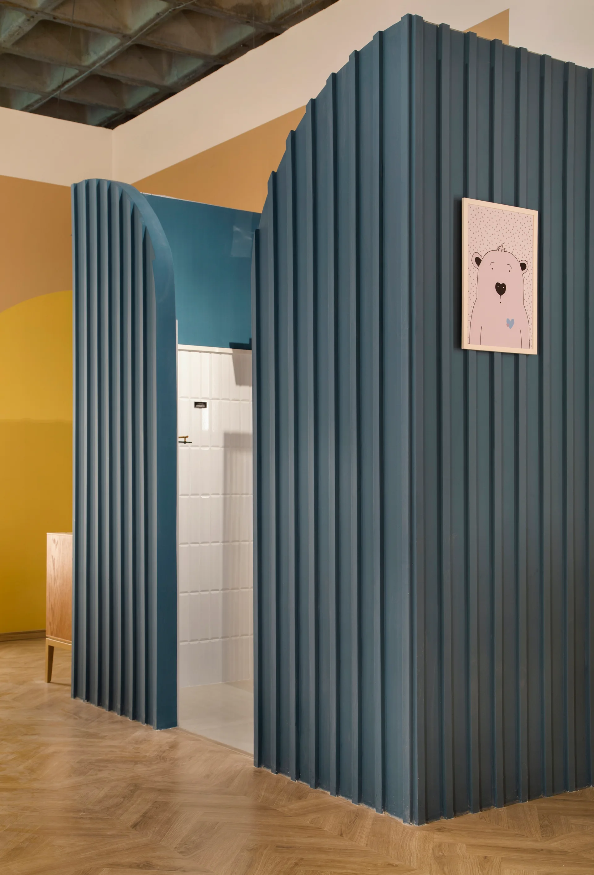 A curved blue fluted partition wall stands on herringbone wood floors next to a white tiled alcove with a framed cartoon bear drawing.