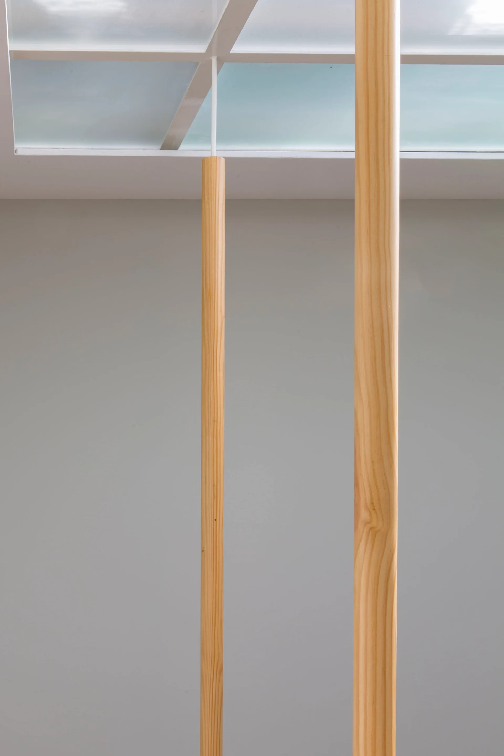 Two slender light wood columns stand vertically in front of a smooth light grey wall beneath a grid-patterned glass skylight.