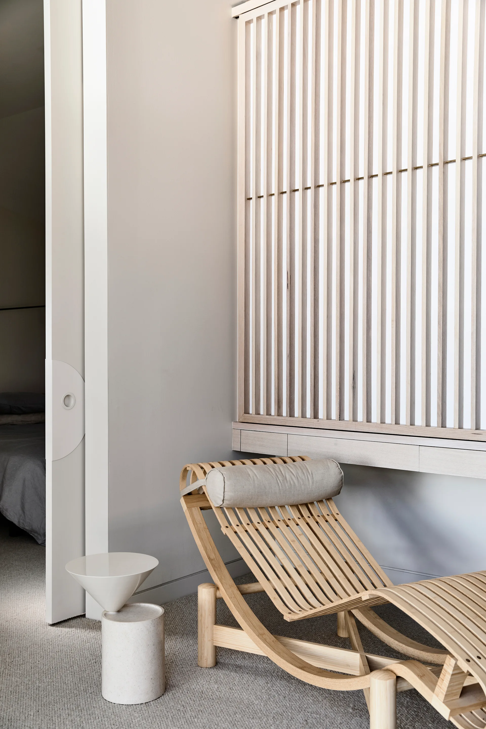 A curved light wood lounge chair with a bolster pillow sits near a tall slatted screen and a sculptural white side table in a minimalist bedroom.