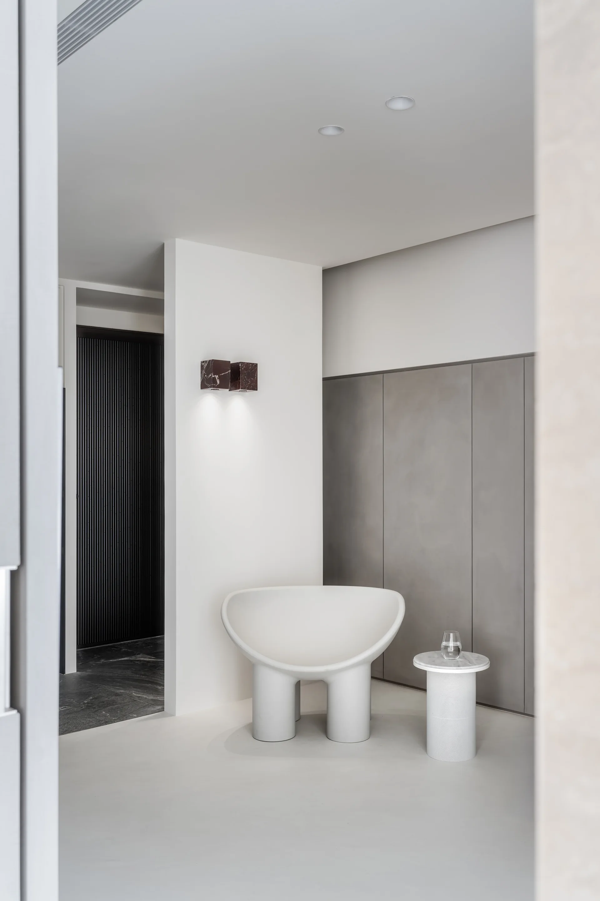 A sculptural white lounge chair and round side table stand in a minimalist hallway corner with grey wall paneling and a dark marble sconce.