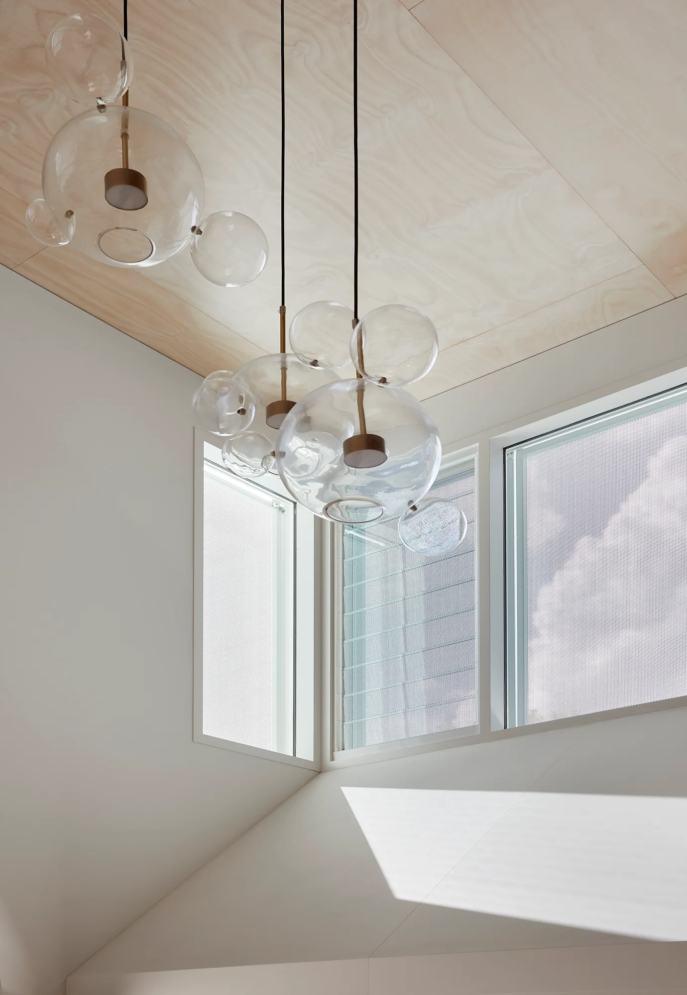Clear bubble glass pendant lights hang from a pale wood-paneled ceiling near high corner windows in a minimalist white room.