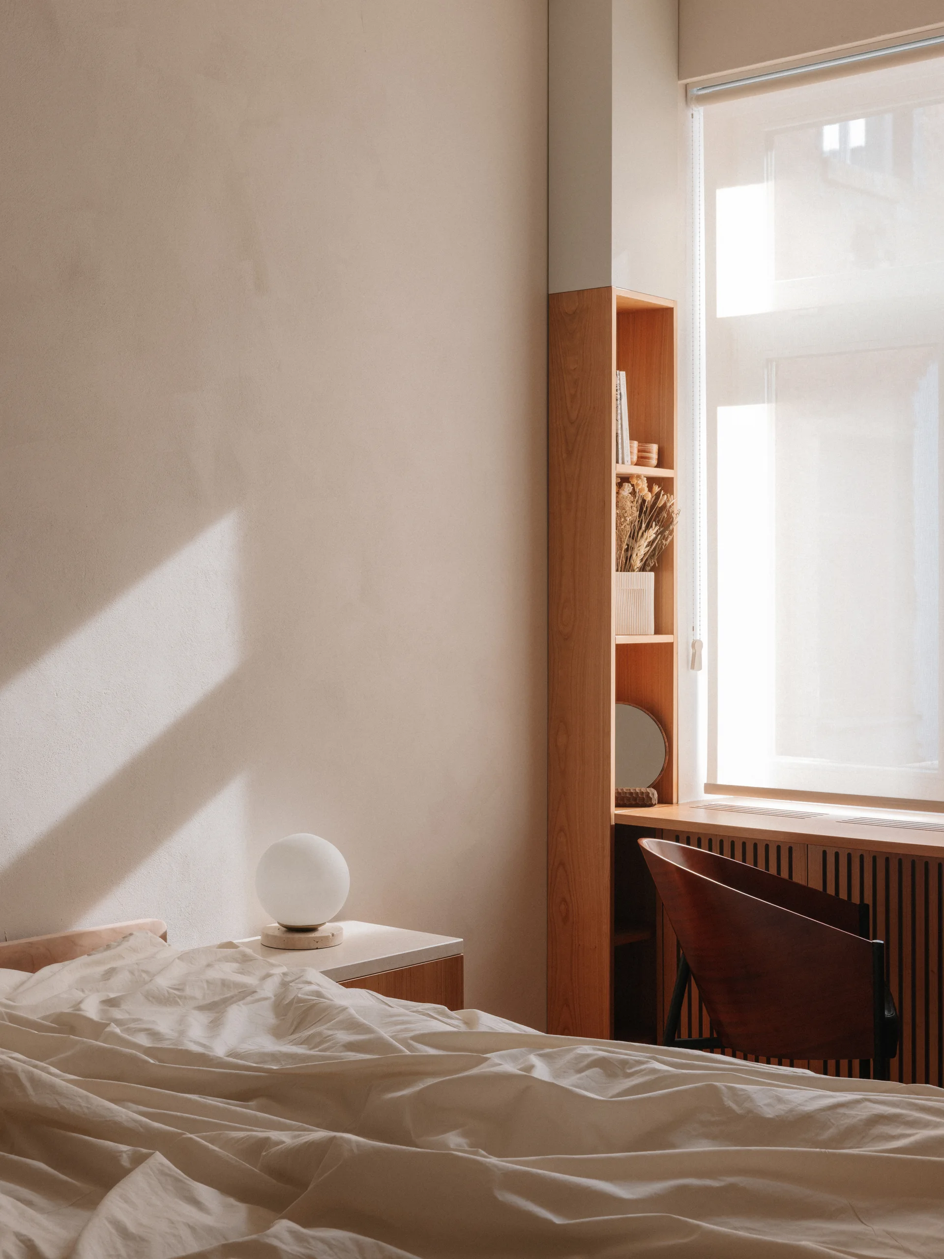 A minimalist bedroom featuring a bed with rumpled linens next to a custom wood shelving unit and desk by a large window.