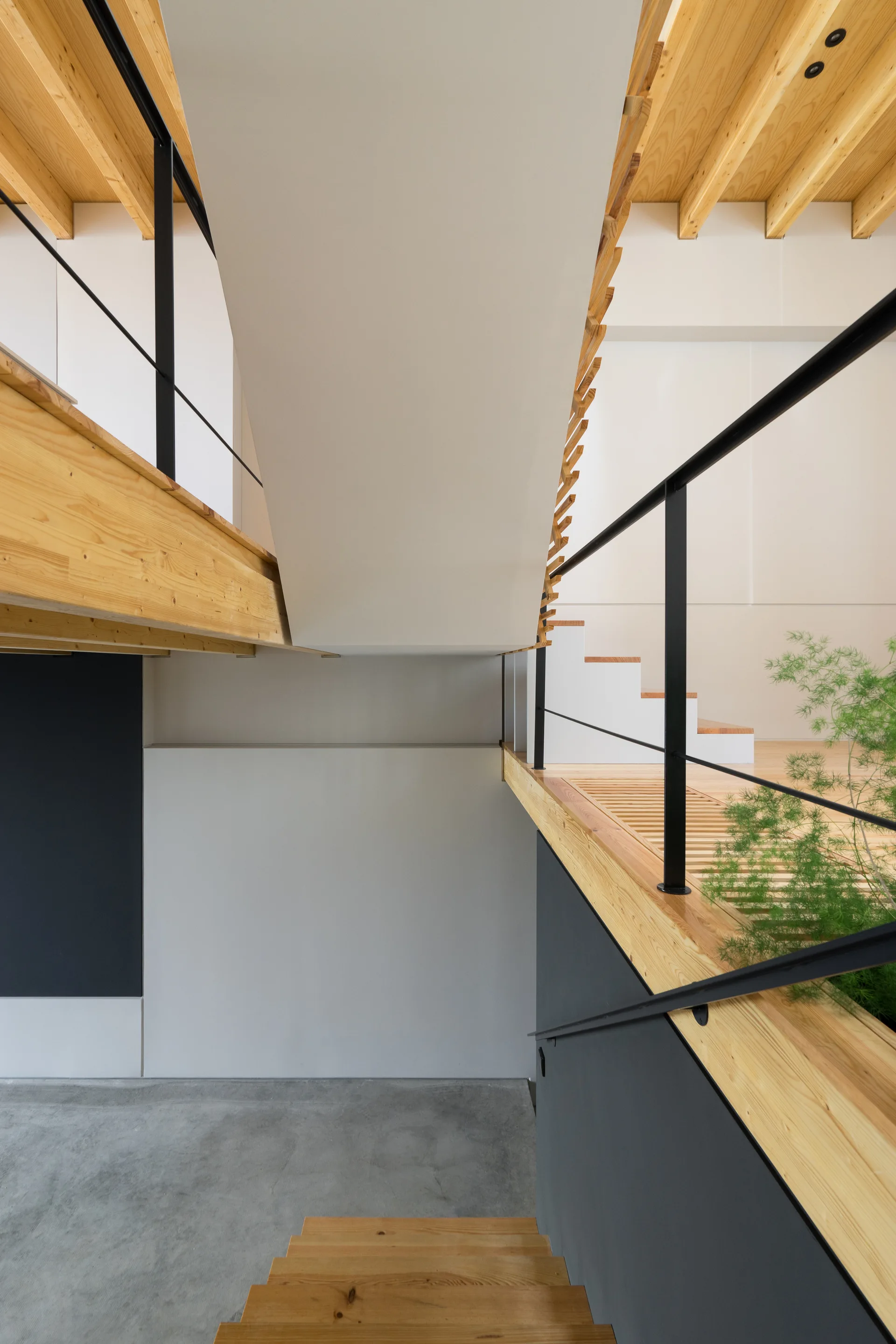 A modern multi-level staircase features natural wood treads, thin black metal handrails, exposed timber ceiling beams, and concrete flooring.