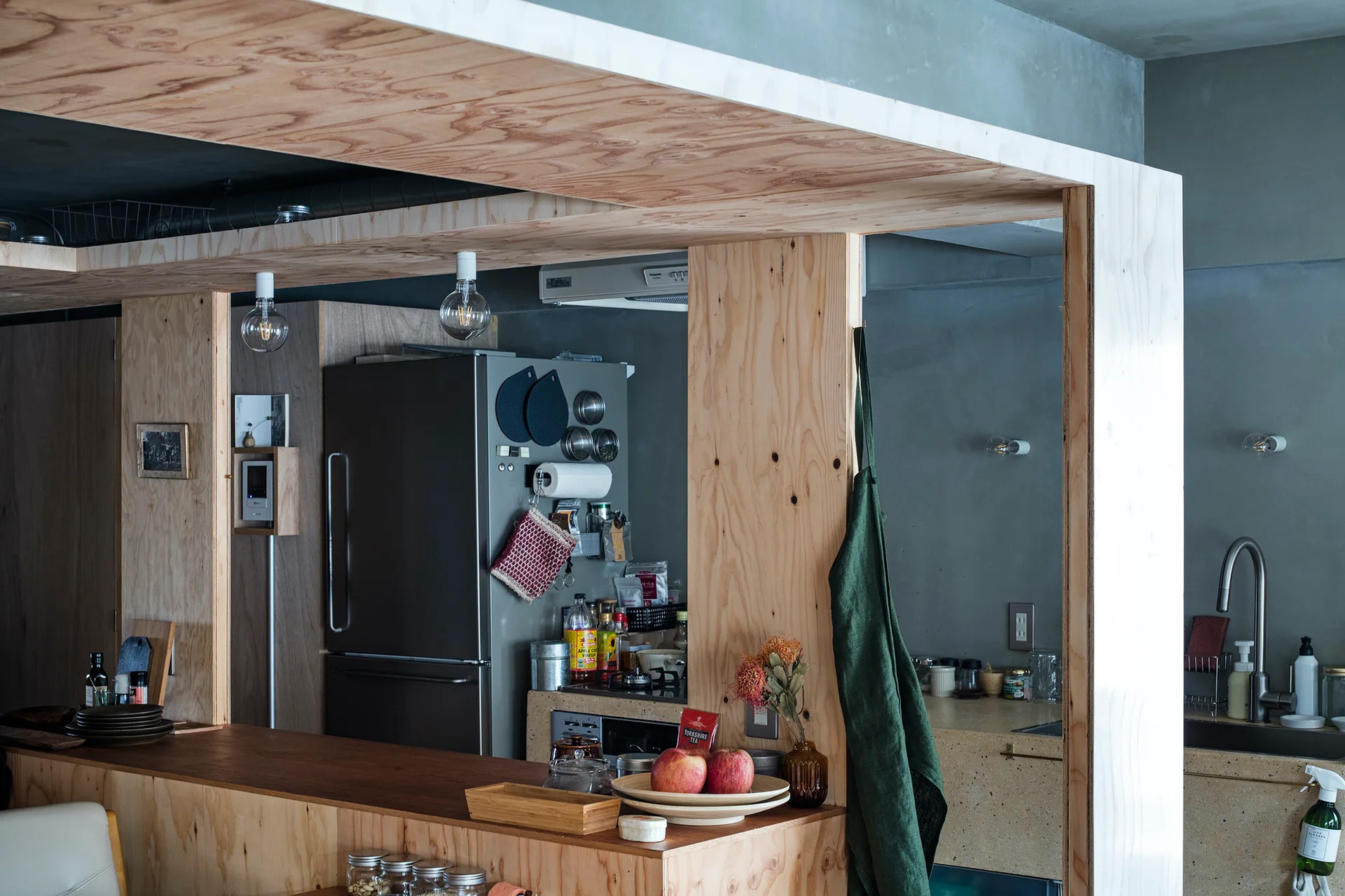 A kitchen interior viewed through a raw plywood partition frame, featuring a wooden counter, a stainless steel refrigerator, and exposed hanging bulbs.