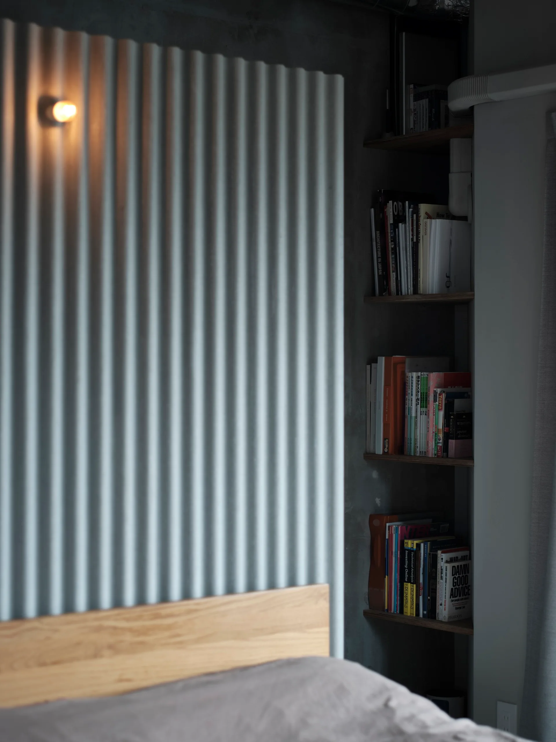 A bedroom corner featuring a grey corrugated wall panel with a small sconce light next to built-in wooden bookshelves set in a concrete niche.