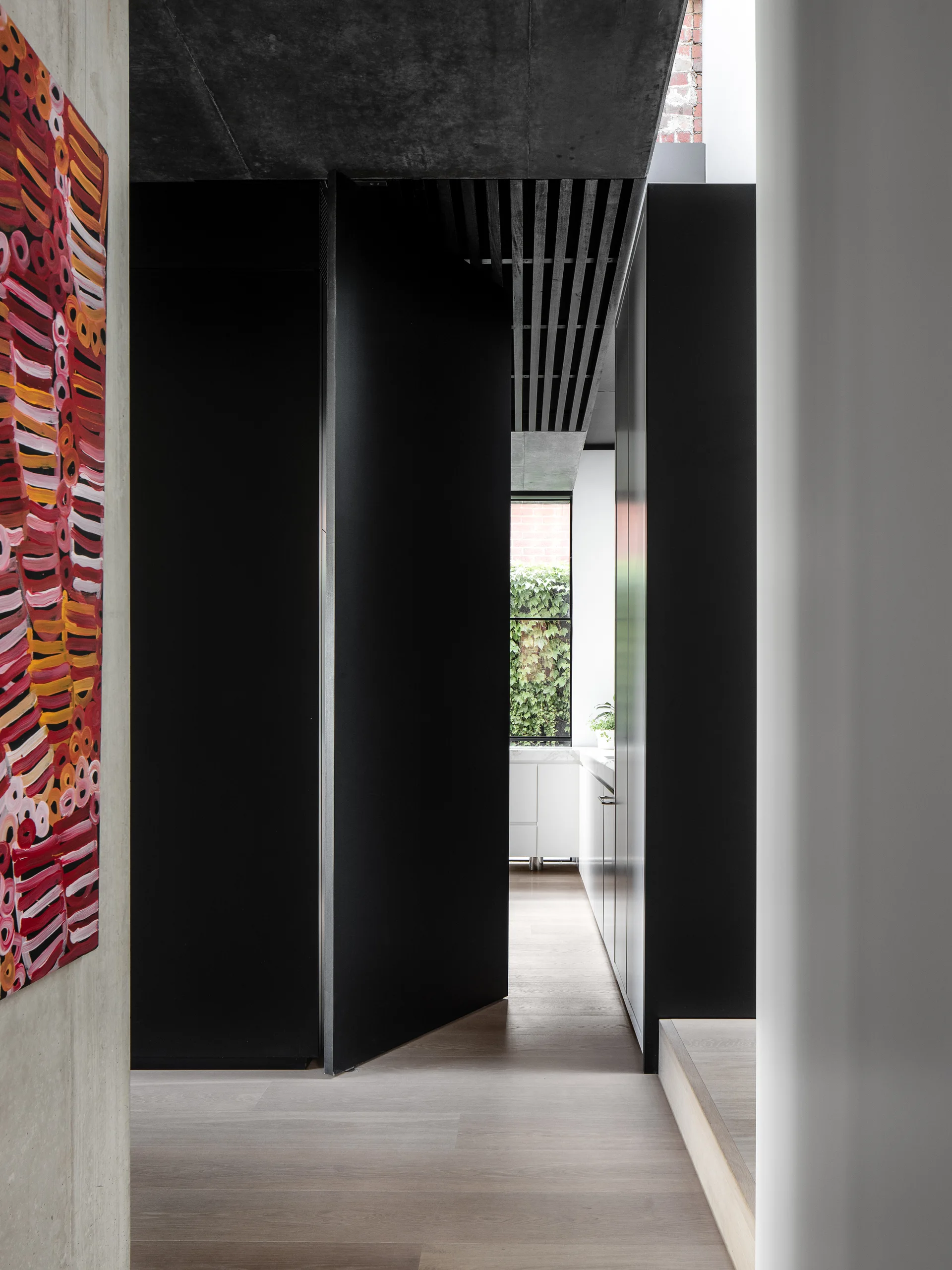 A minimalist hallway with a large matte black pivot door, raw concrete surfaces, light wood floors, and a bright abstract painting on the wall.