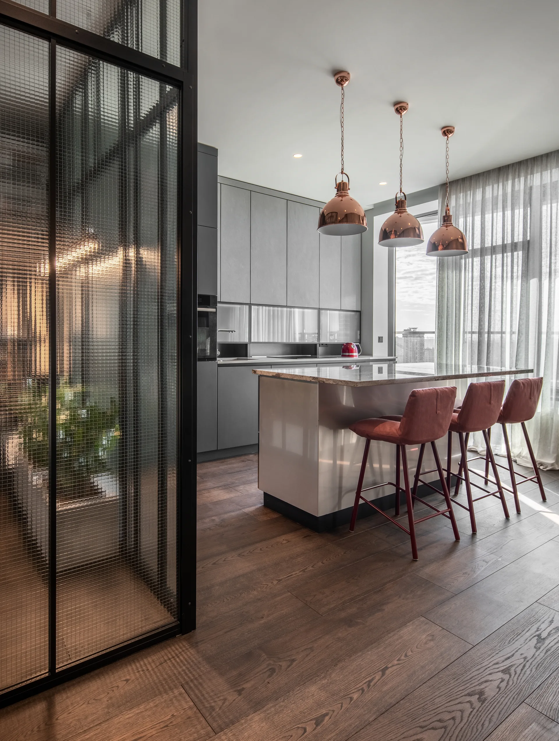 A contemporary kitchen featuring a central island with red stools, copper pendant lights, and a wire-mesh glass partition.