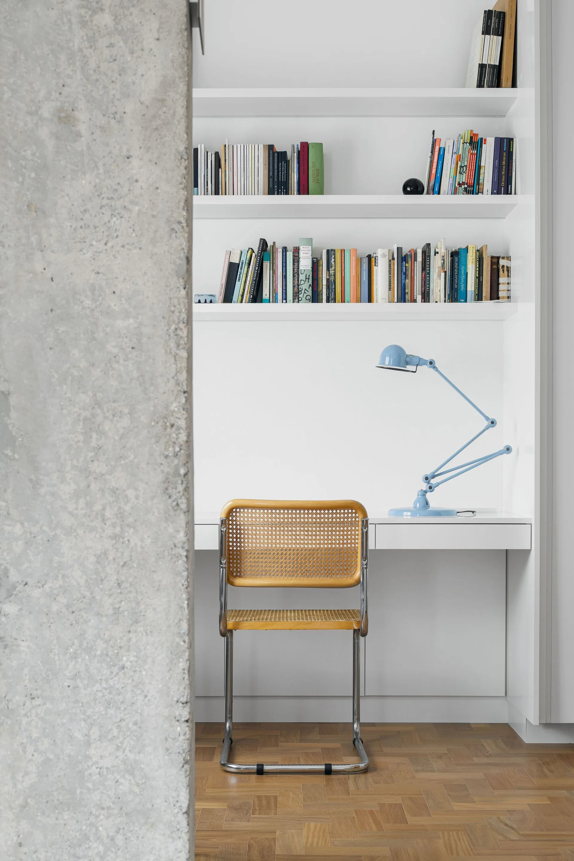 A minimalist built-in white desk with bookshelves features a woven cane chair, a light blue task lamp, and parquet flooring next to a concrete column.