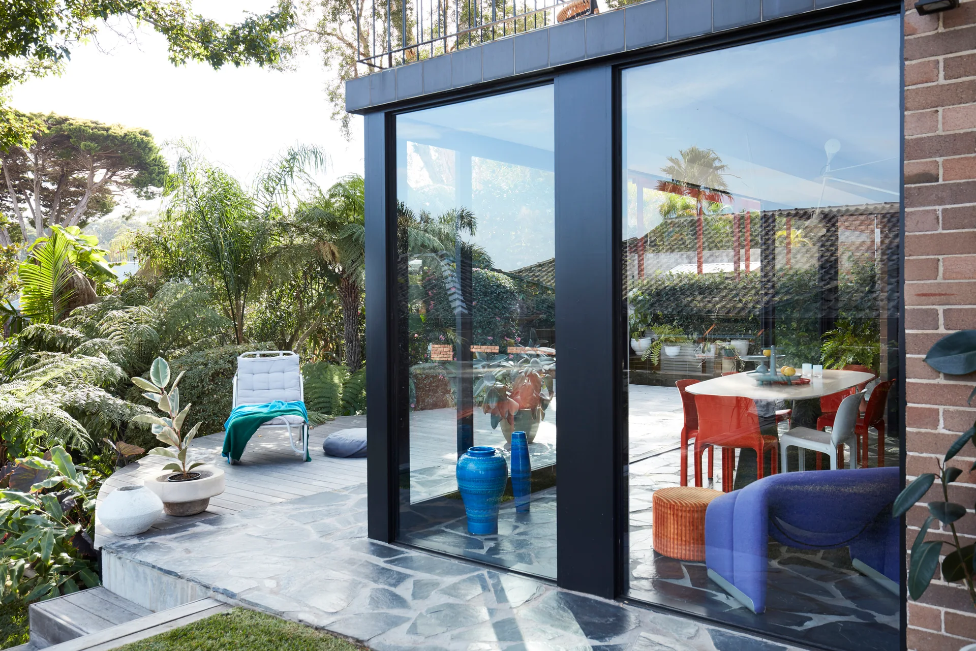 A flagstone patio with a white lounge chair sits outside large black-framed glass sliding doors revealing a colorful dining and living room interior.