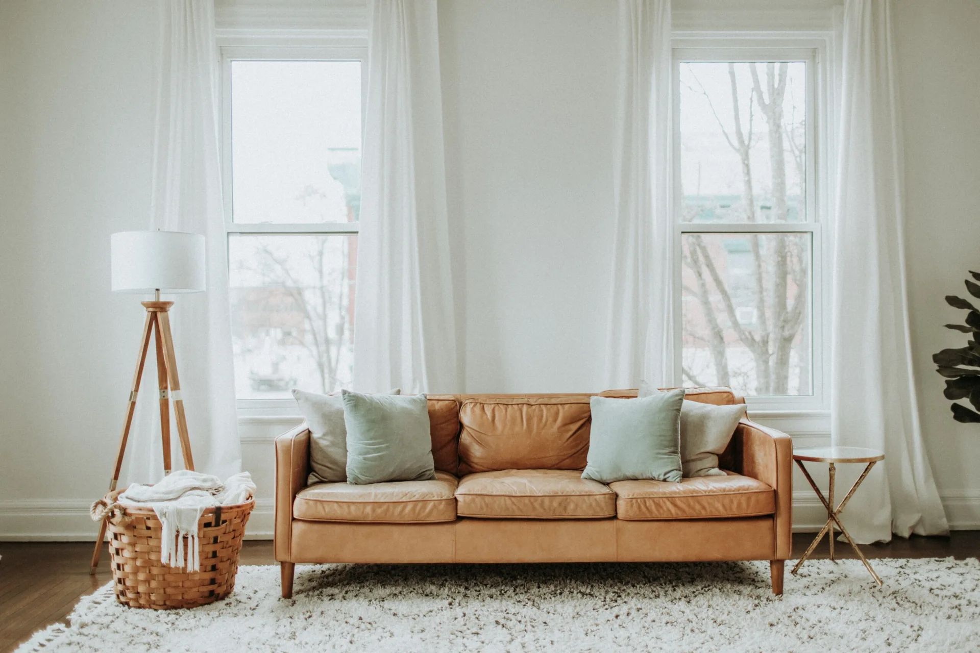 A tan leather sofa sits on a textured white shag rug between two tall windows with white drapes in a bright living room.