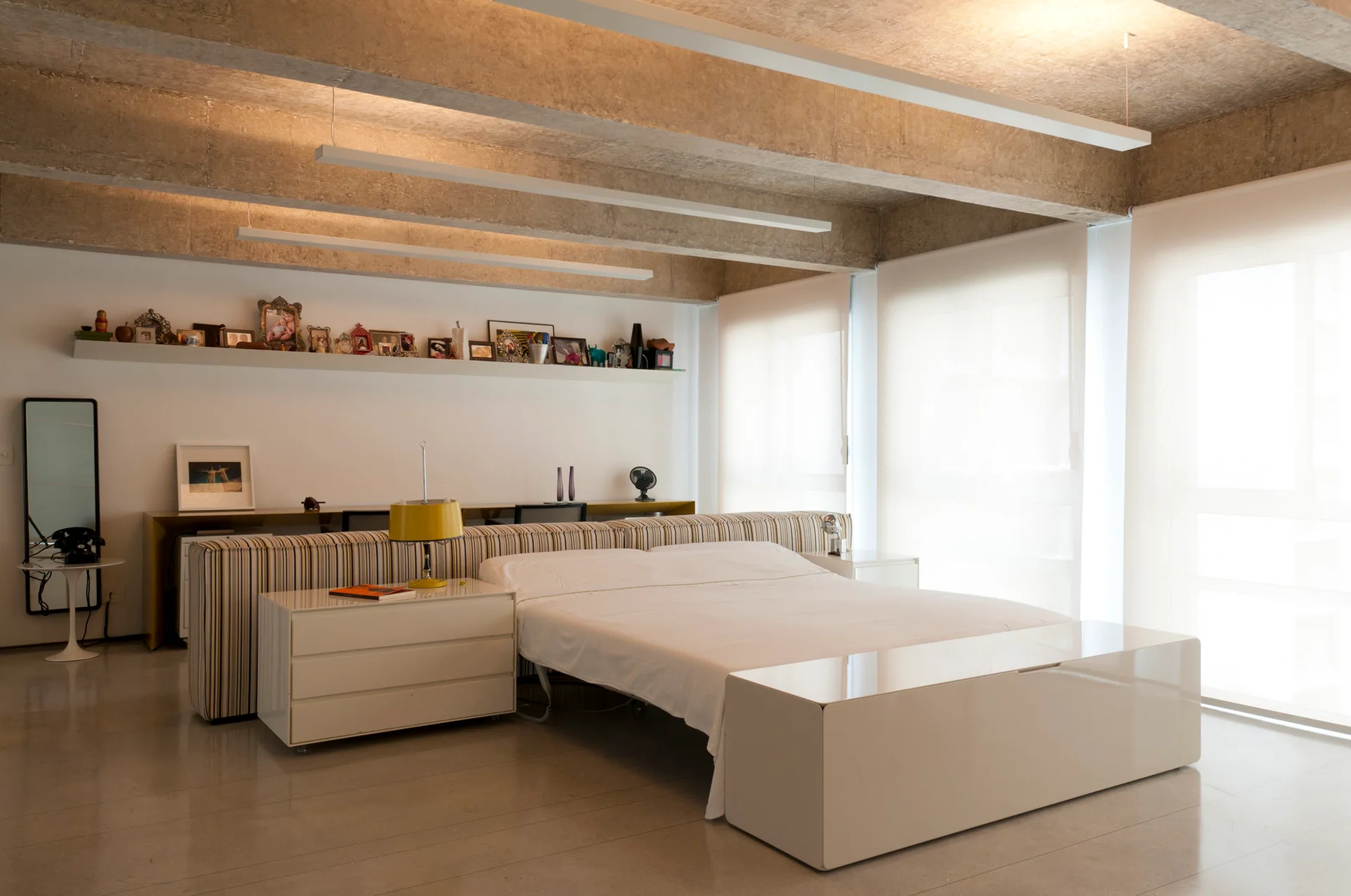 A bedroom with exposed concrete ceiling beams, a low white bed, a striped headboard divider, and a desk behind it.