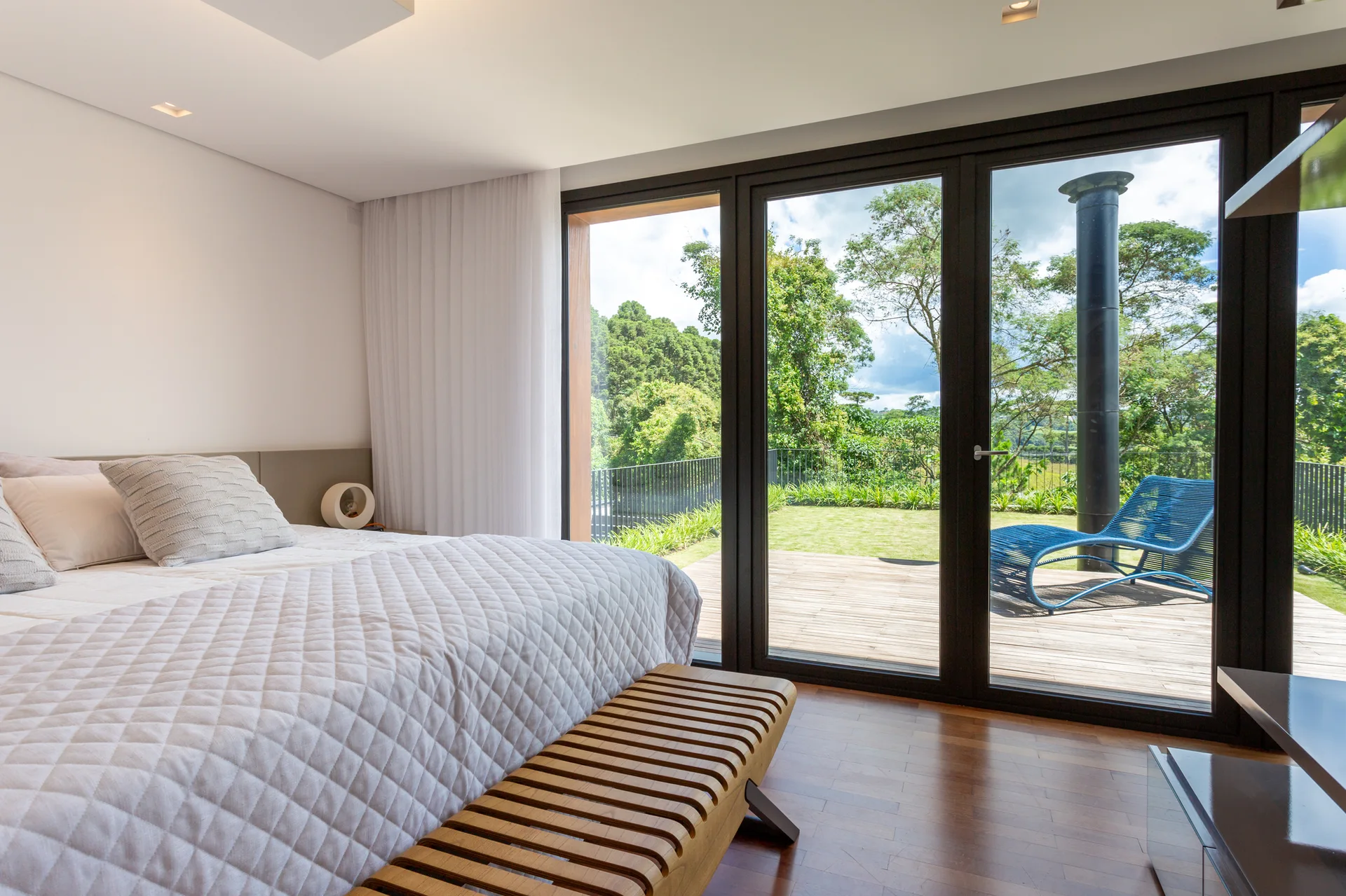 A modern bedroom with a quilted bed and a slatted wood bench looking out to a garden deck through large glass doors.