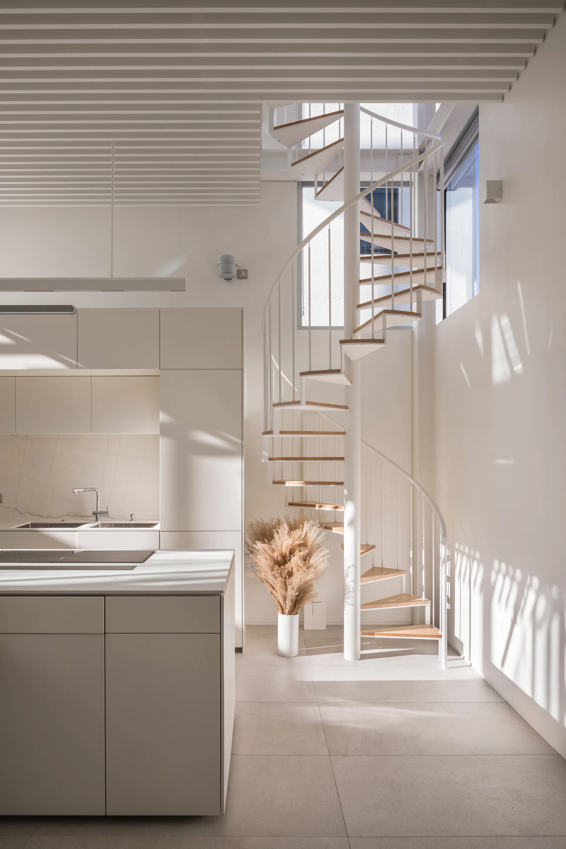 A modern minimalist kitchen features off-white cabinetry, a stone island, and a white metal spiral staircase with wood treads under a slatted ceiling.