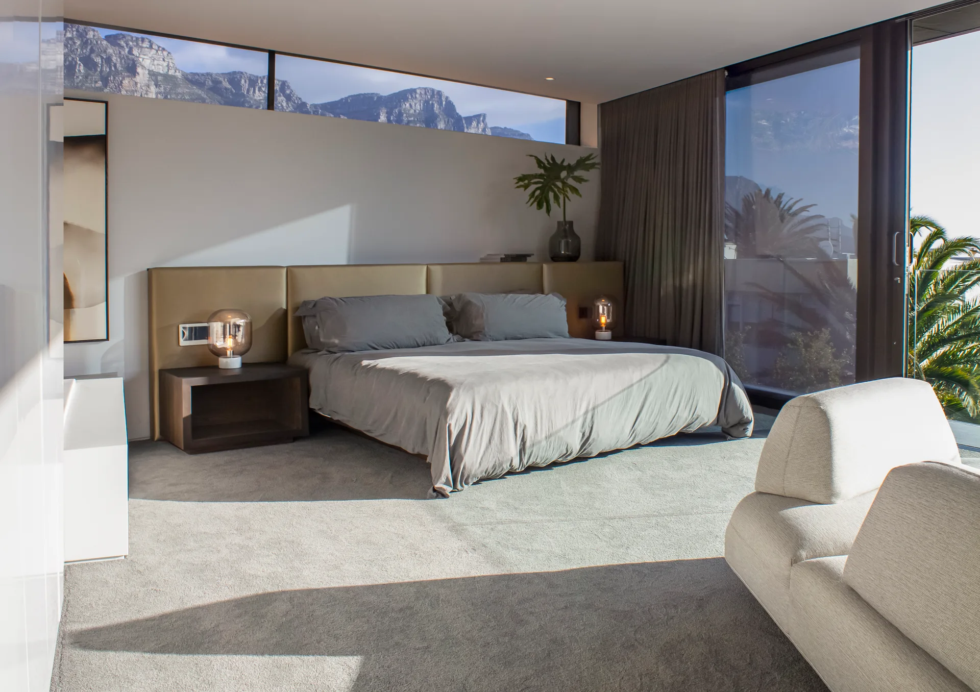 A contemporary bedroom features a king bed with a long beige headboard, dark wood nightstands, and a high horizontal window showing mountain views.
