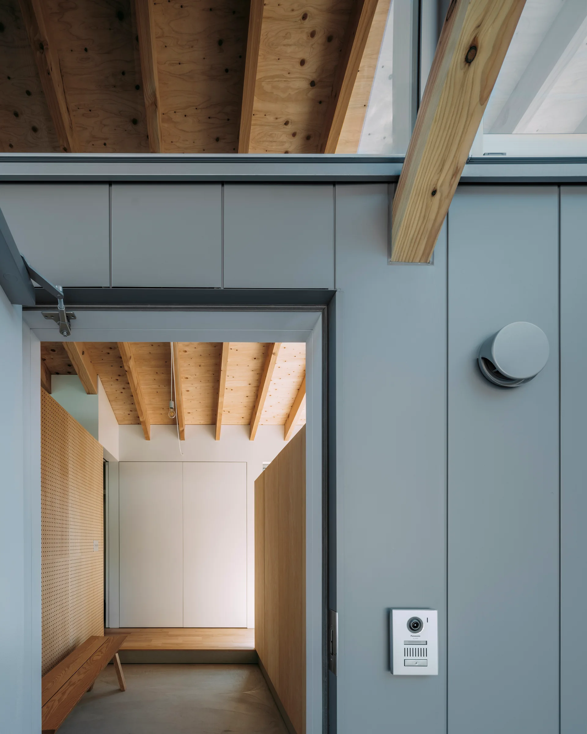 An entryway doorway looking into a space with a wood bench, pegboard wall, and exposed timber ceiling rafters.