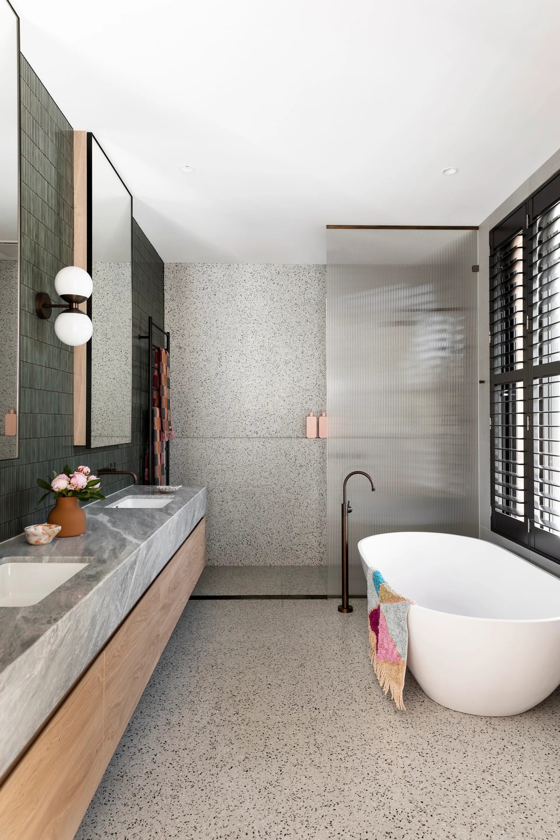 A modern bathroom features a wood vanity with a grey stone countertop, a white freestanding bathtub, a fluted glass screen, and speckled terrazzo flooring.