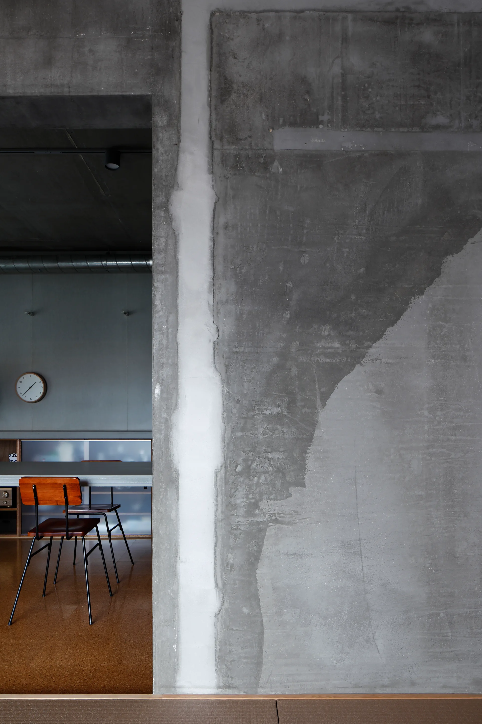 An opening in a raw gray concrete wall reveals a minimalist dining room with wood-and-metal chairs, a gray table, and a wall clock.