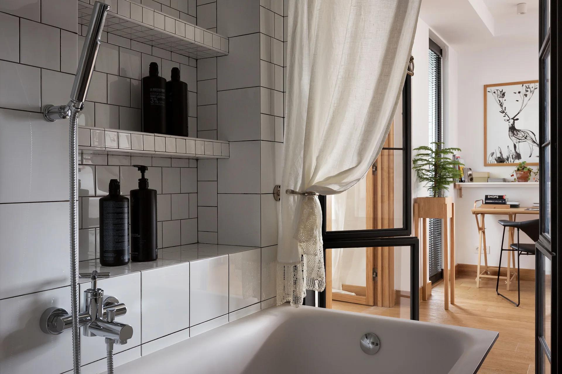 A white tiled bathroom with a chrome shower head and tiled shelves overlooks an open-plan home office with wooden flooring and a desk.