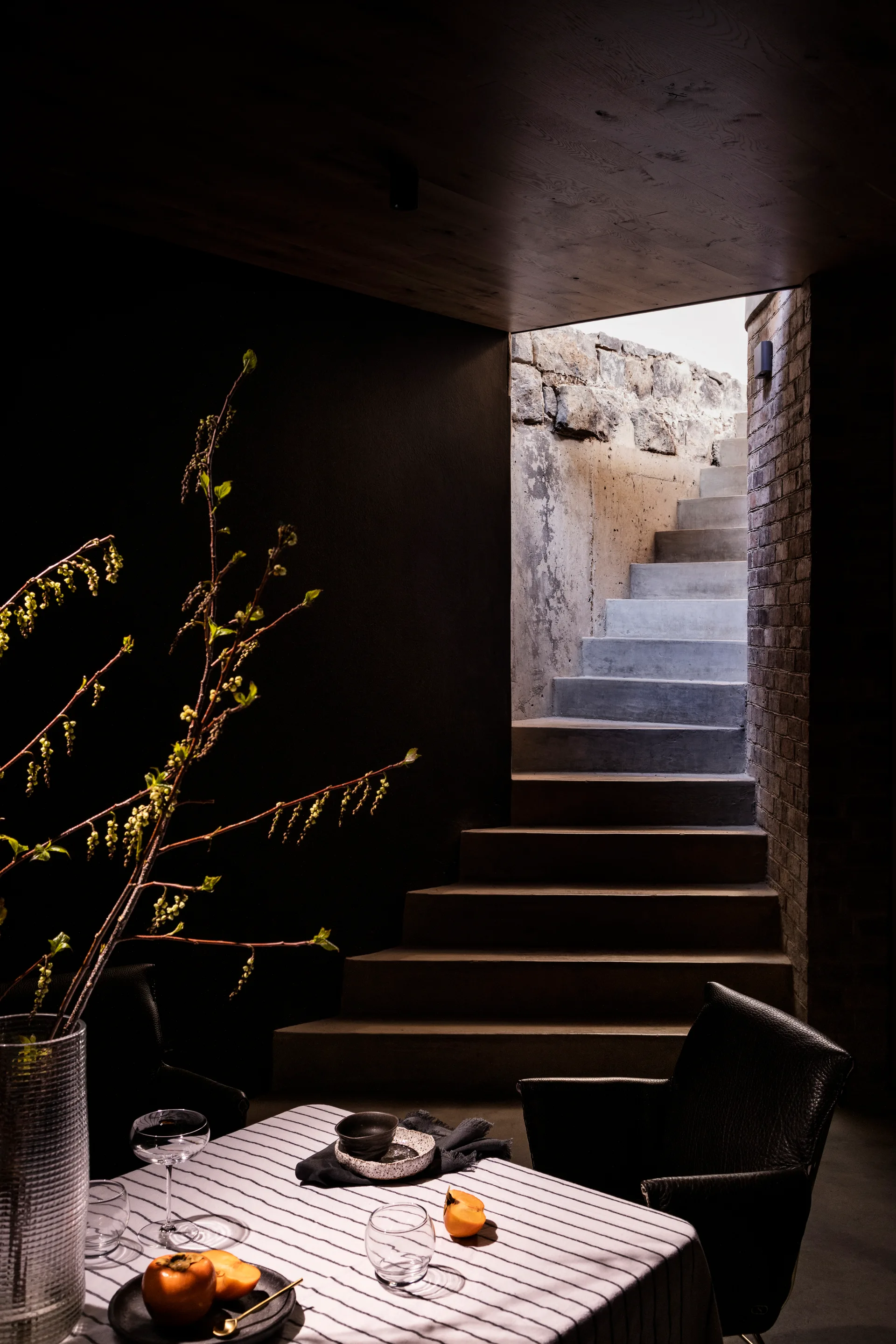 A dimly lit dining table with a striped tablecloth sits before a concrete staircase illuminated by natural light.