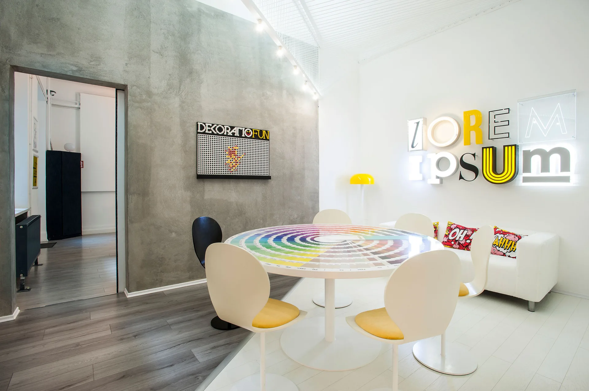 A modern dining area features a circular color wheel table, white modern chairs with yellow cushions, and a textured concrete wall.