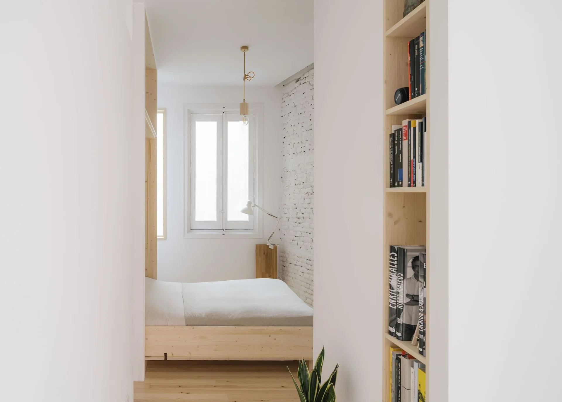 A minimalist bedroom viewed through a doorway, featuring a light wood bed frame, a white painted brick wall, and a built-in wooden bookshelf.