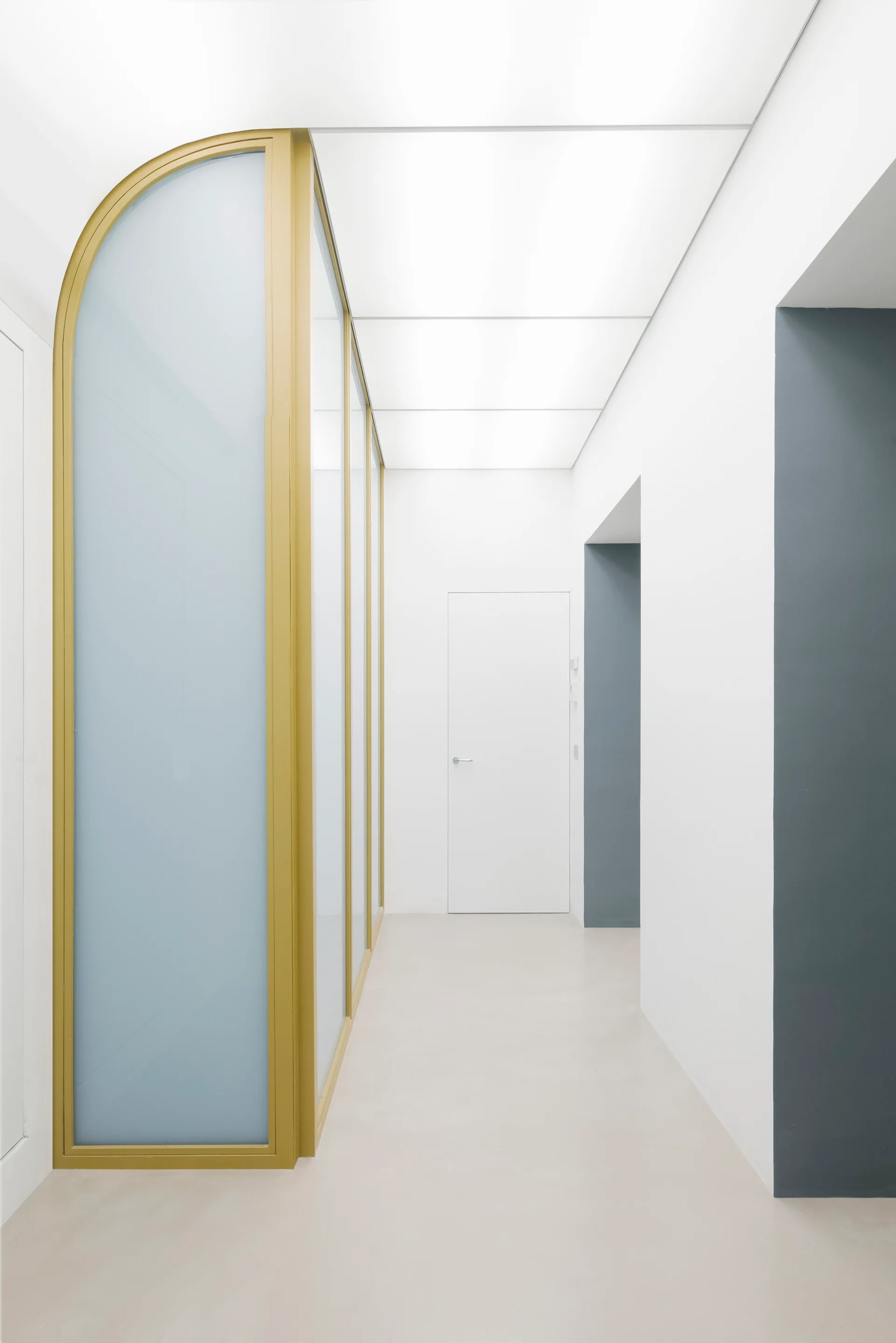 A minimalist hallway with a gold-framed arched glass partition, white walls, and a recessed grey doorway.