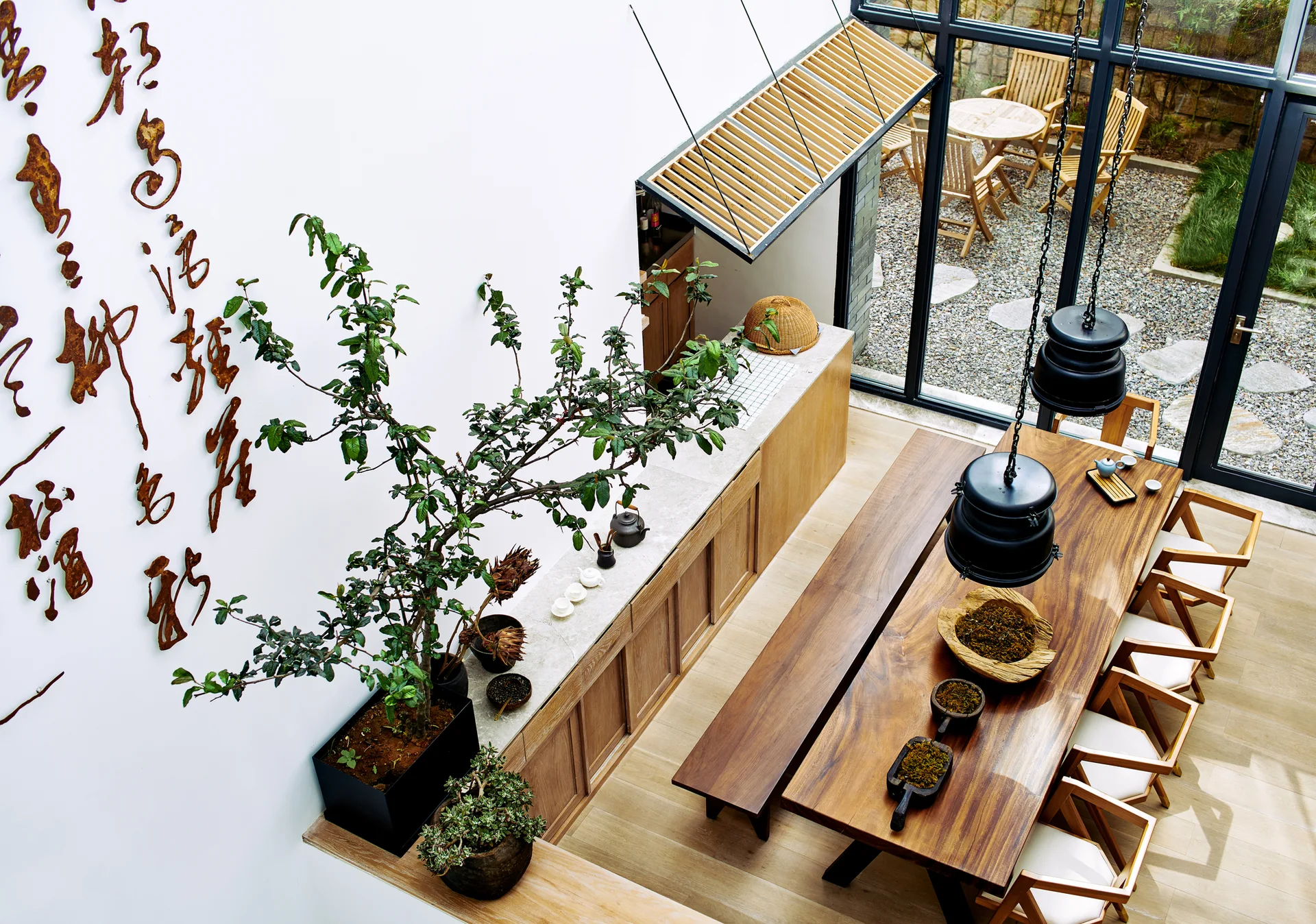 High-angle view of a modern dining room with a long wooden table, bench, chairs, black pendant lights, and calligraphy on a white wall.