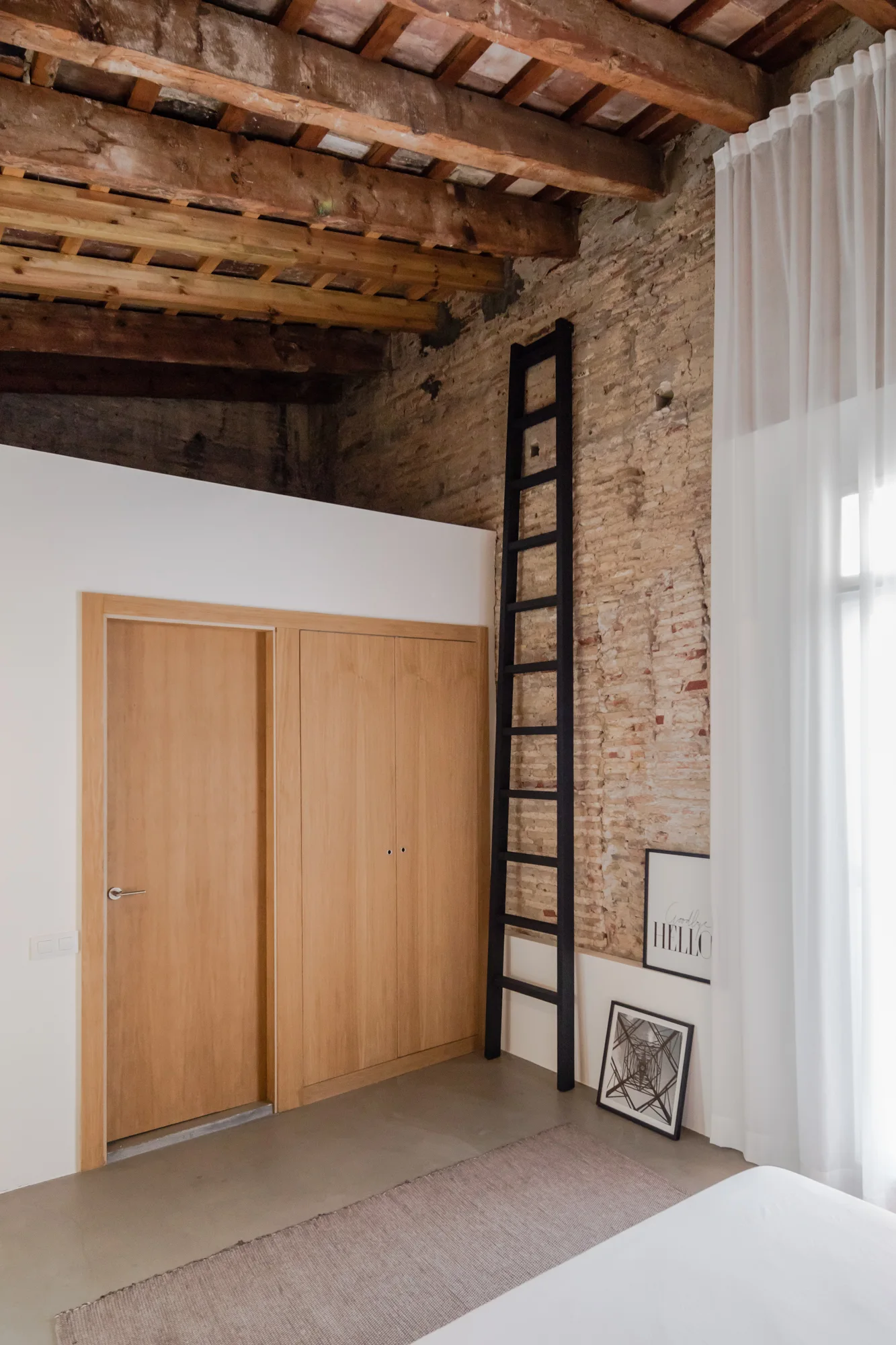 A bedroom corner featuring an exposed brick wall, timber-beamed ceiling, wood wardrobe, and a tall black ladder leaning next to sheer white curtains.