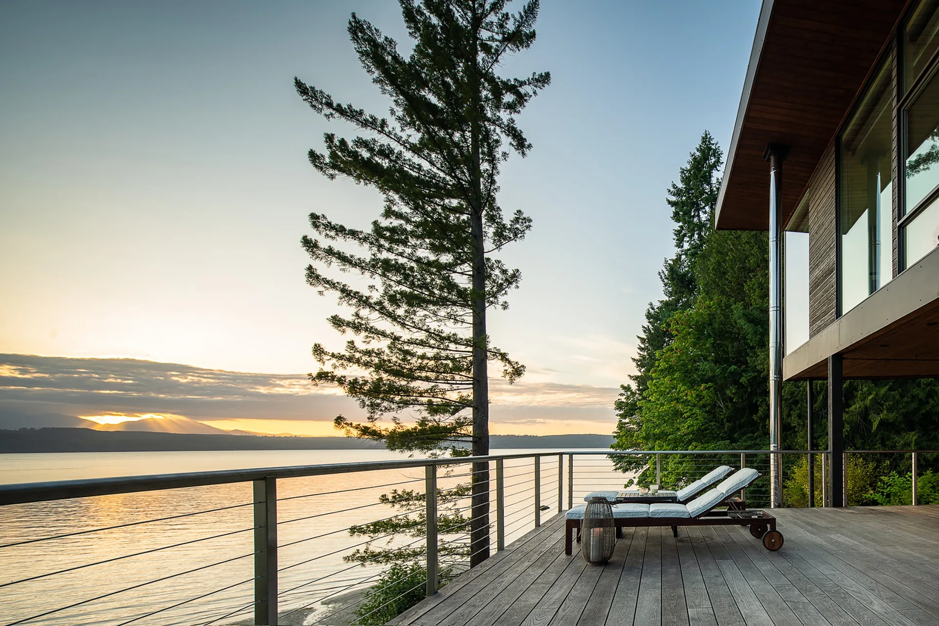 Two wooden lounge chairs with white cushions sit on a spacious wood deck overlooking a calm lake at sunrise behind a metal cable railing.