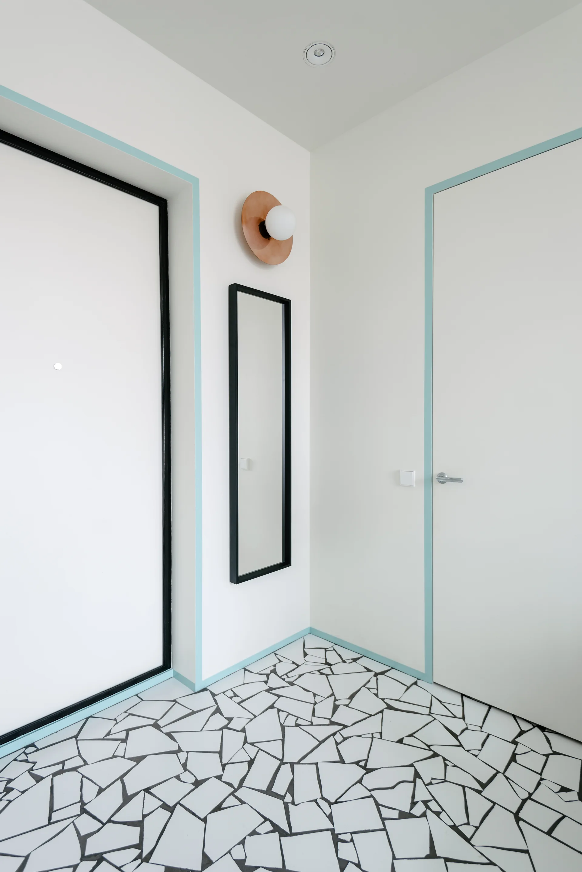 A minimalist entryway with a black and white fractured mosaic floor, white doors outlined in light blue trim, and a narrow wall mirror.