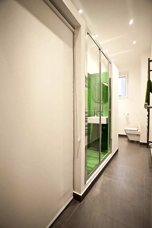 A narrow modern bathroom with dark grey floor tiles, white walls, and a glass shower enclosure lined with green mosaic tiles.