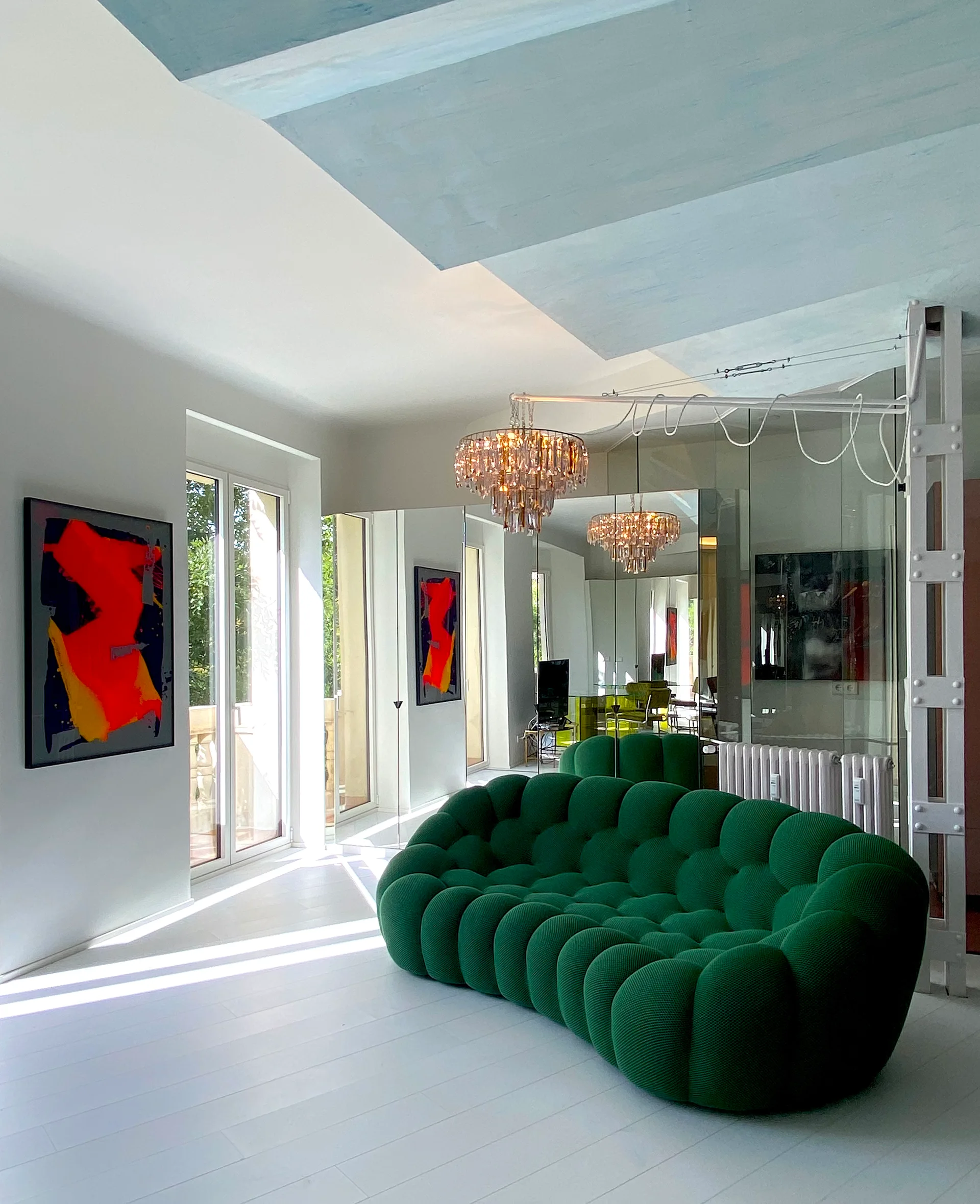 A bright living room featuring a large green tufted sofa positioned in front of a mirrored wall reflecting a crystal chandelier.