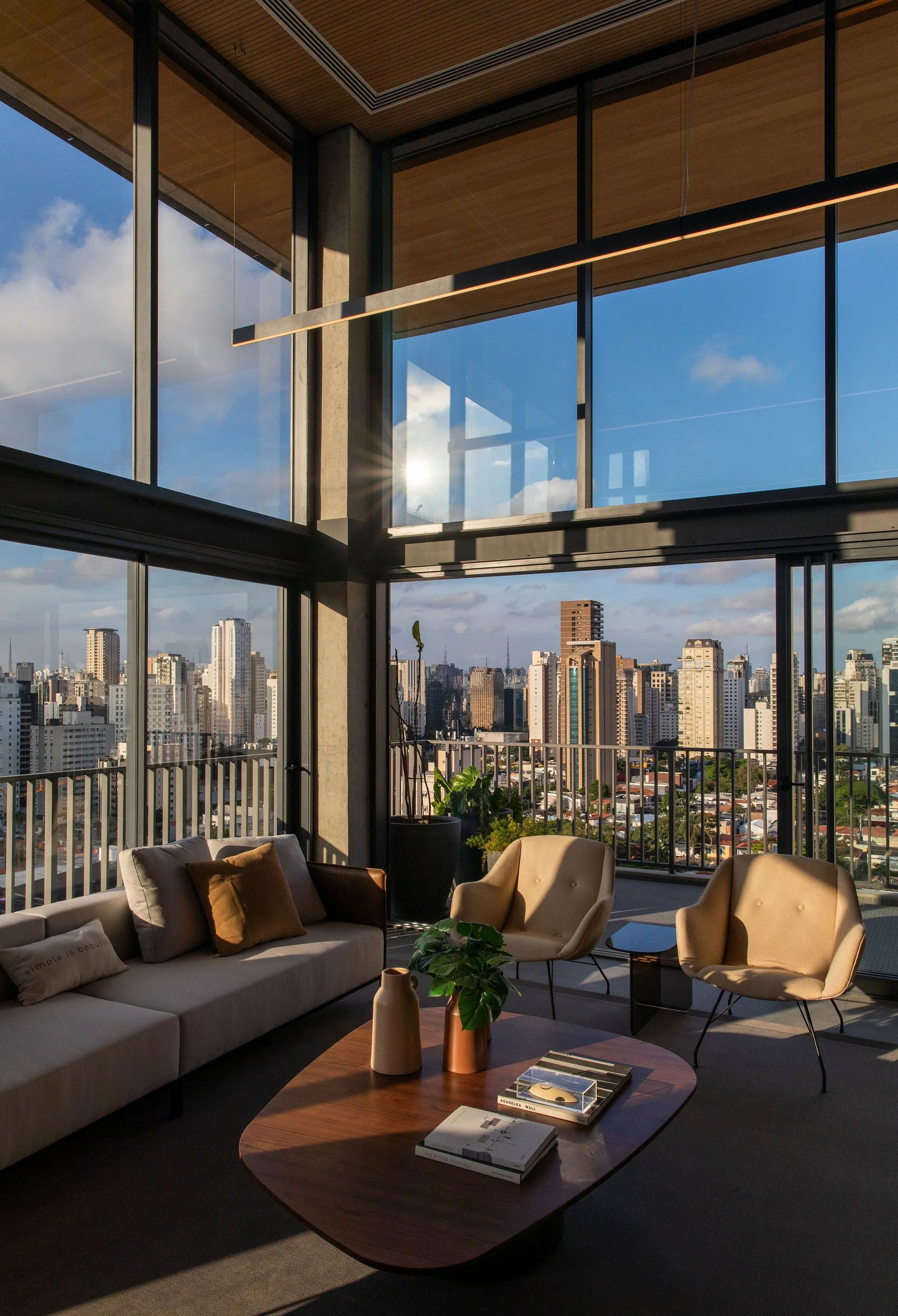 A modern living room features a beige sofa, tan armchairs, a wood coffee table, and large glass walls looking out onto a city skyline.