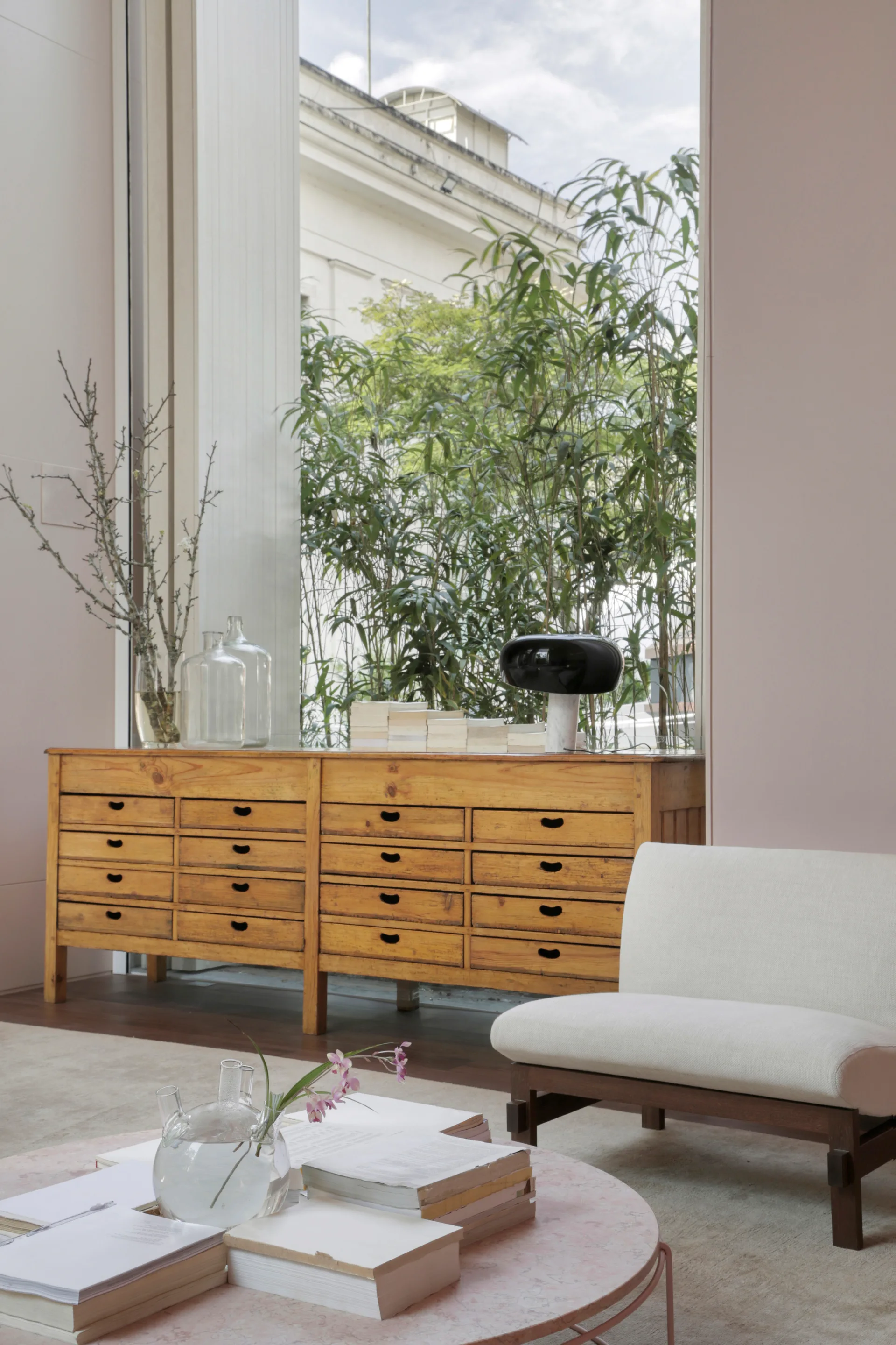 A pale pink living room featuring a multi-drawer wooden chest of drawers, a low-profile white armchair, and a stone-topped coffee table by a large window.